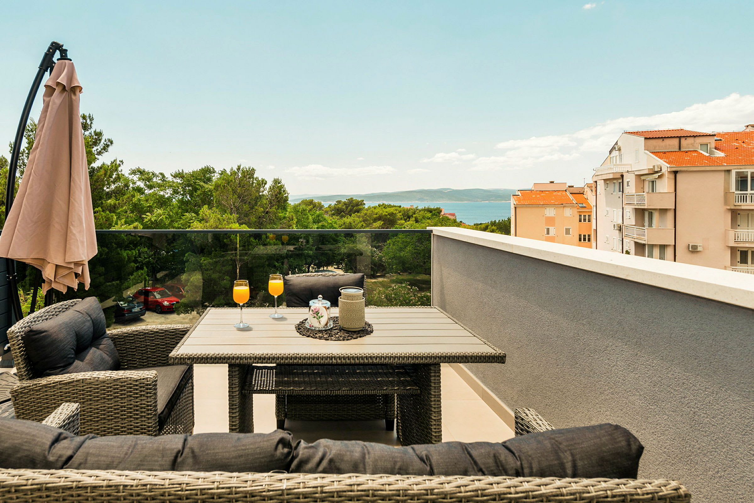 Terrasse med bord og stole, orange drikke i glas, parasol og udsigt over træer, bygninger og hav. (AI-genereret tekst)