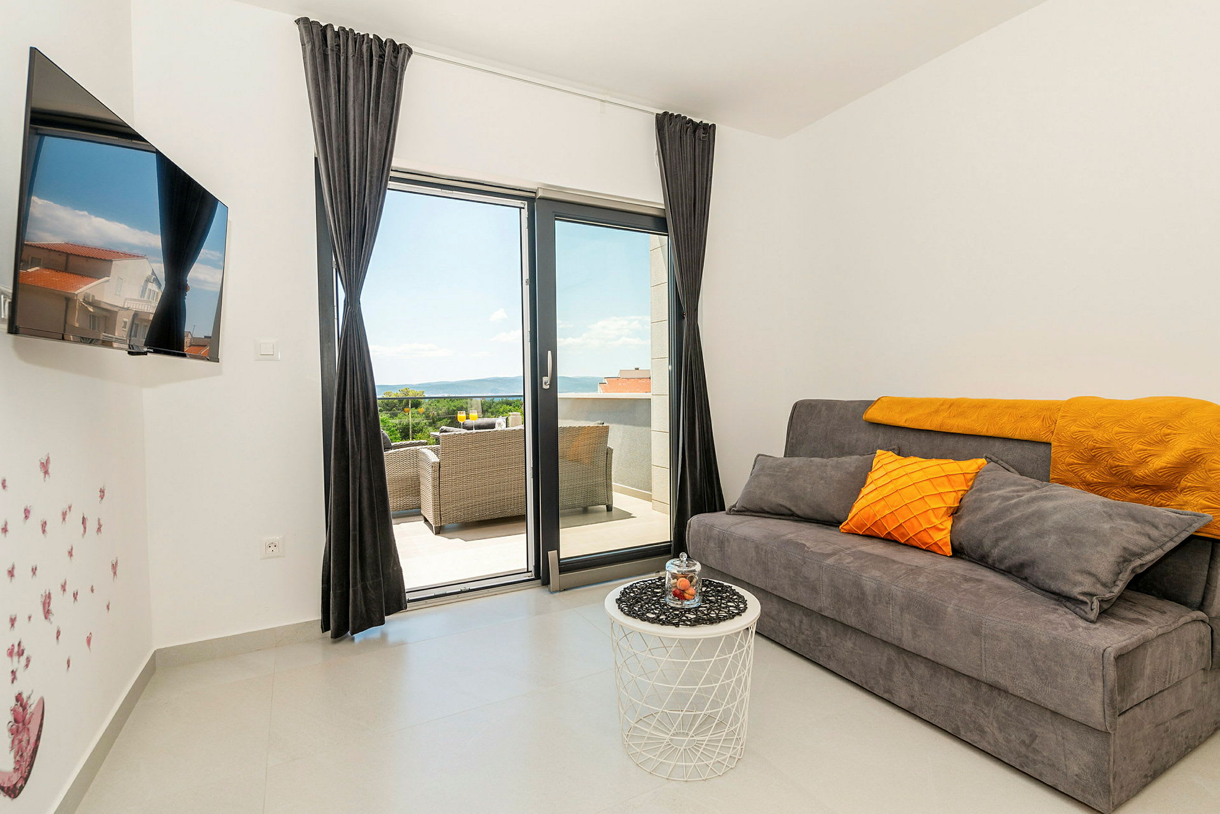 En moderne stue med en grå sofa, orange puder, et hvidt bord og en balkon med udsigt. (AI-genereret tekst)