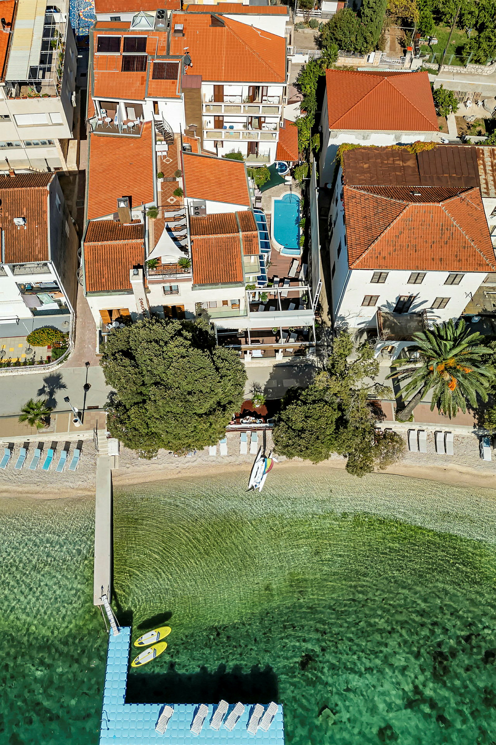 Luftfoto af en bygning med tagterrasse, en pool og en bro med kajakker ved en strand. (AI-genereret tekst)