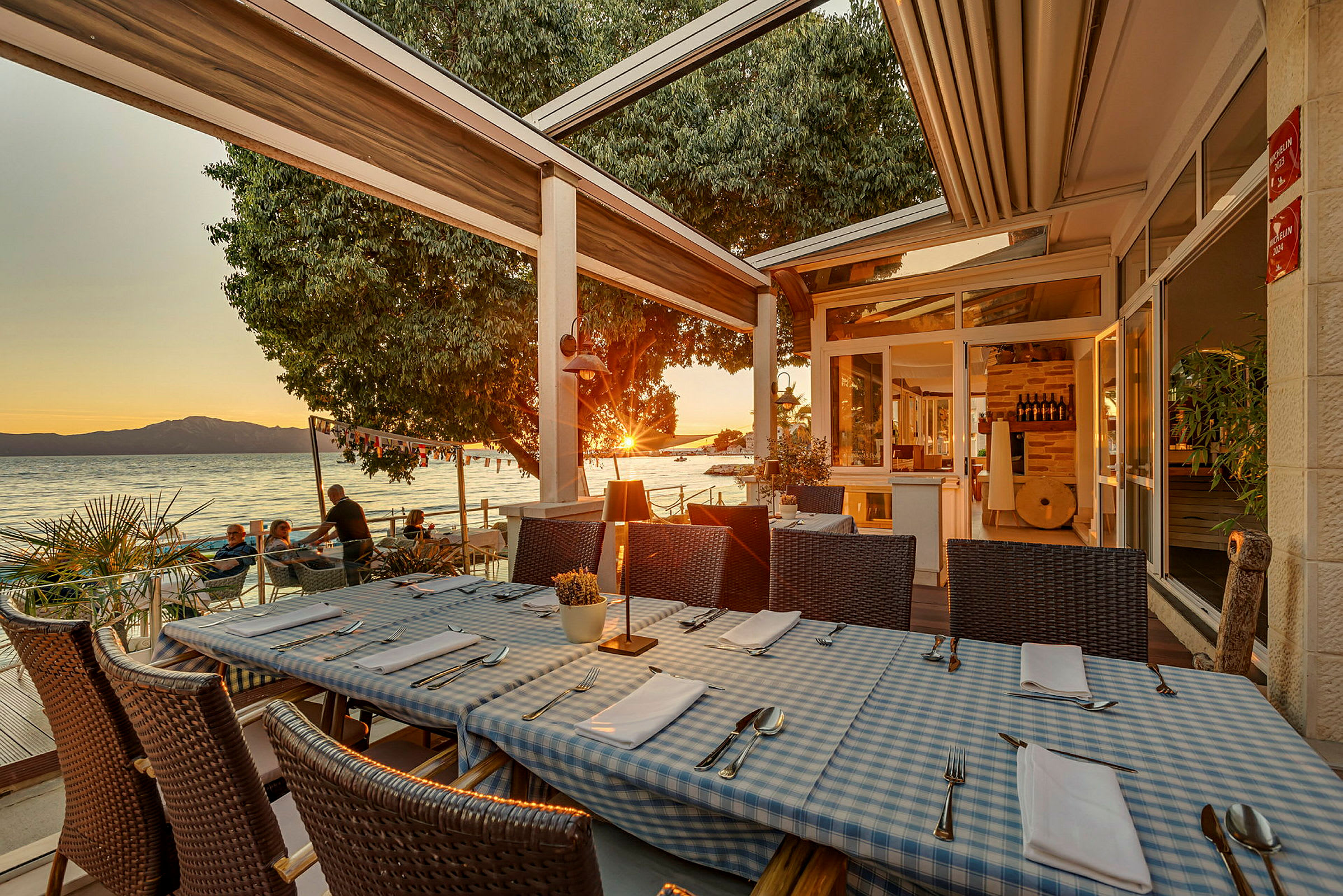 Restaurantborde med blå- og hvidternede duge på en terrasse ved havet under solnedgangen. (AI-genereret tekst)