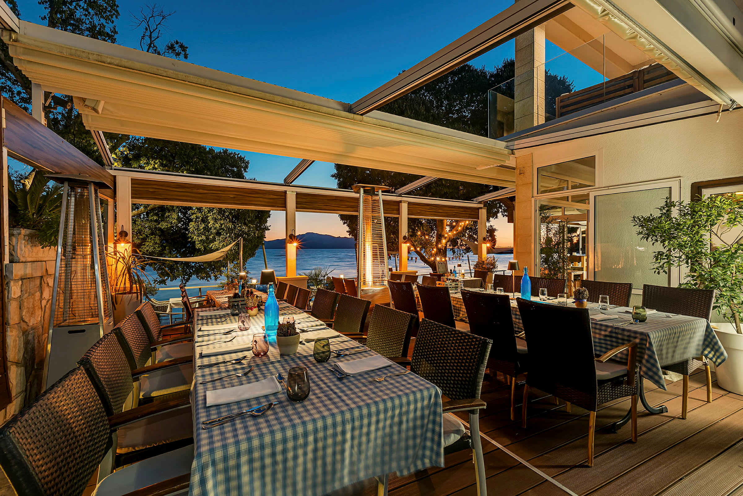 Billede af en udendørsrestaurant med opdækkede borde og udsigt til havet. (AI-genereret tekst)