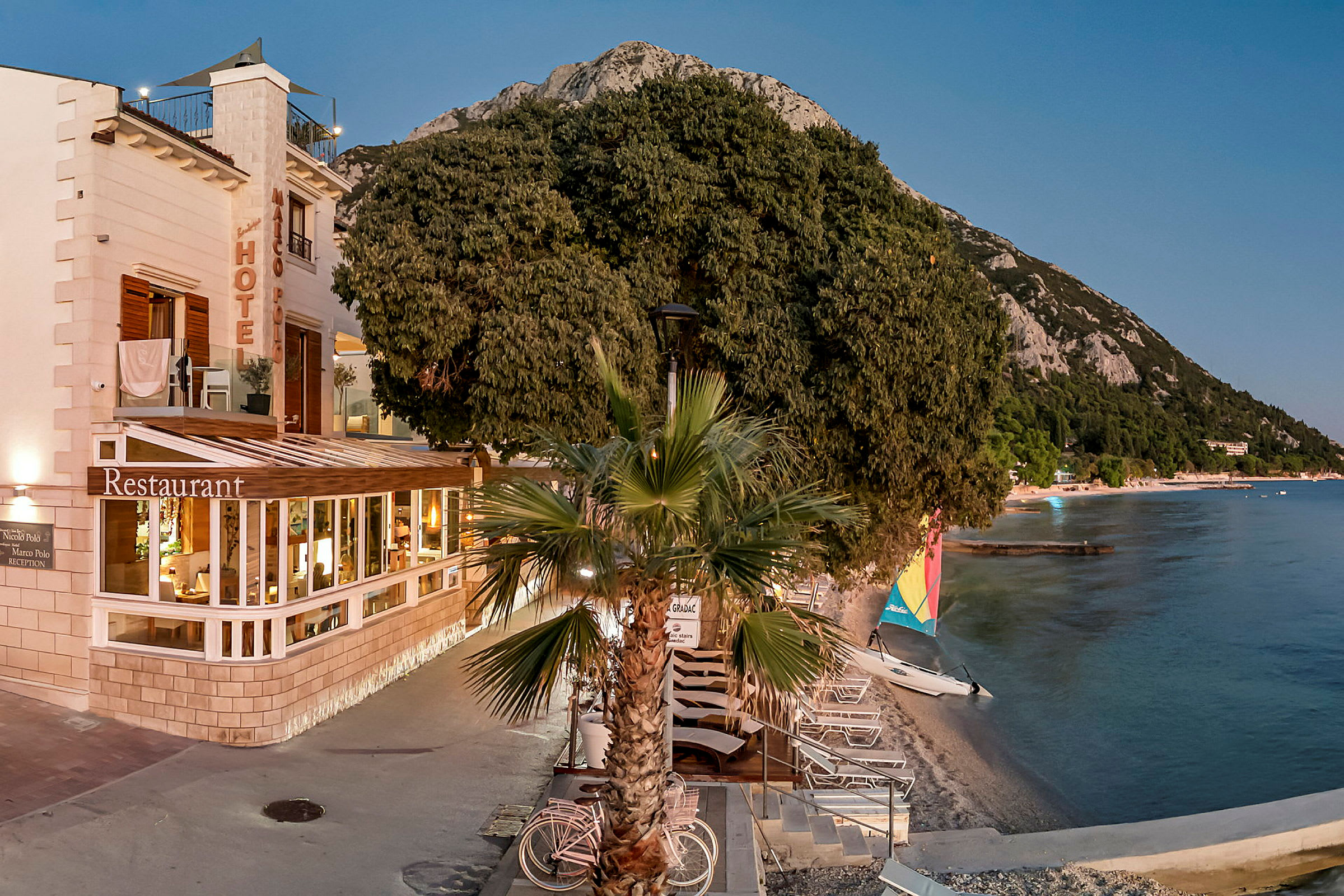 Billede af et hotel ved stranden, med en restaurant og liggestole langs vandkanten. (AI-genereret tekst)