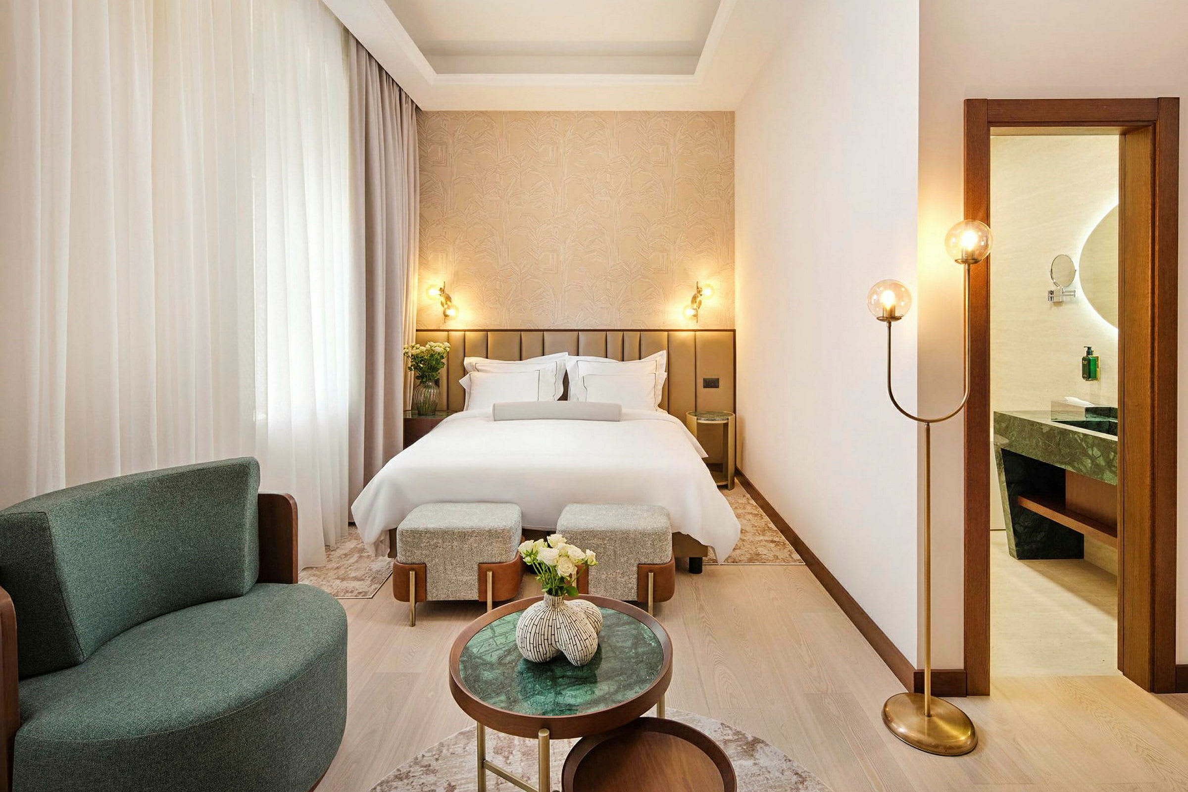 Et elegant hotelværelse med en redt seng, lænestol og små borde. Et badeværelse ses gennem en åben dør. (AI-genereret tekst)