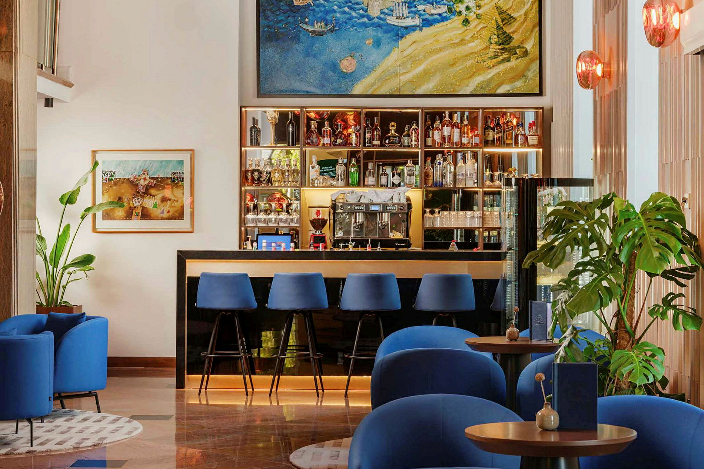 En hotelbar med blå stole ved disken samt blå lænestole foran små borde. Vægsdekorationer og planter findes i baggrunden. (AI-genereret tekst)