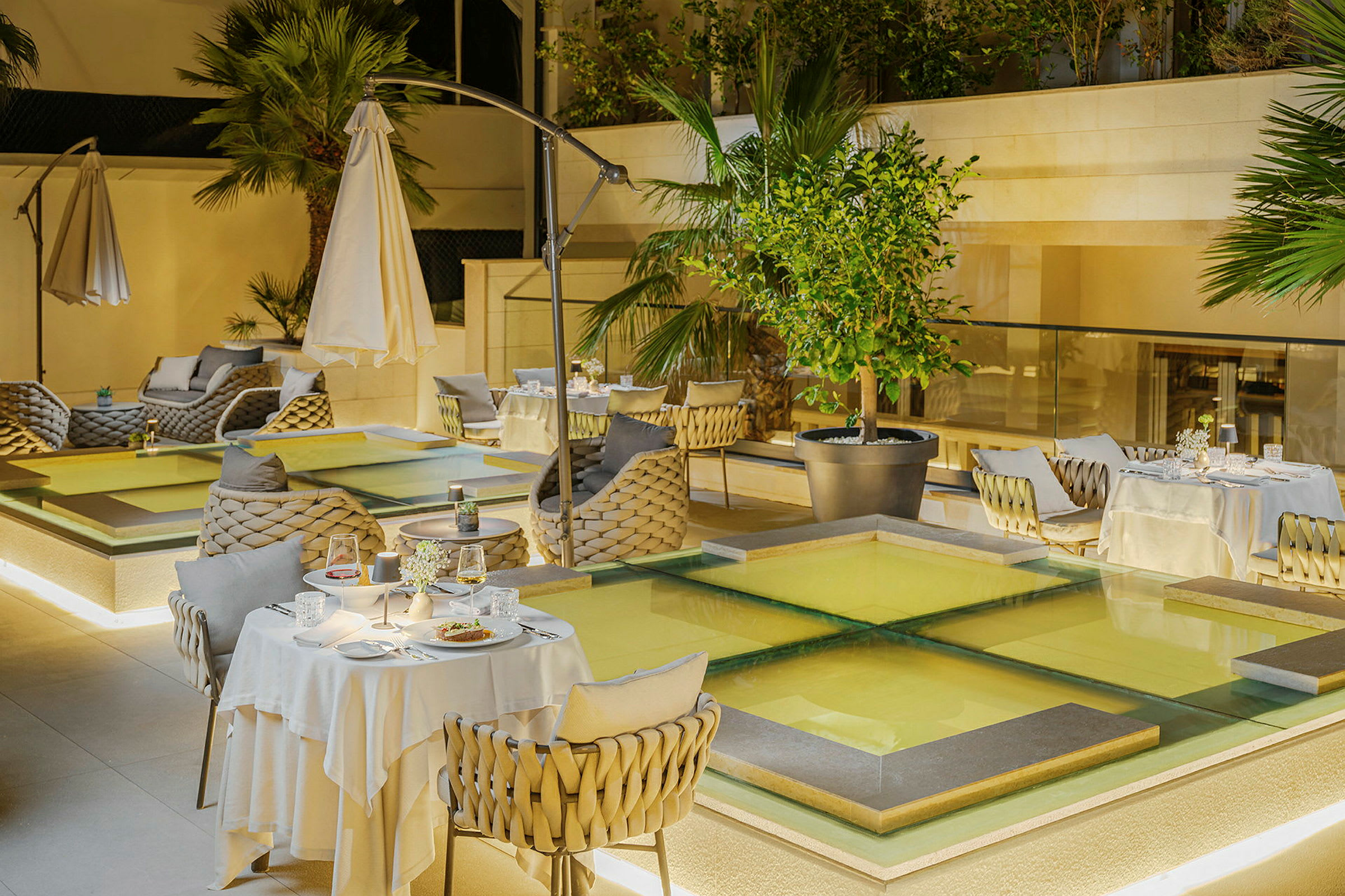 En elegant udendørsrestaurant med dækkede borde, stole i flettet design og omkringliggende beplantning. (AI-genereret tekst)