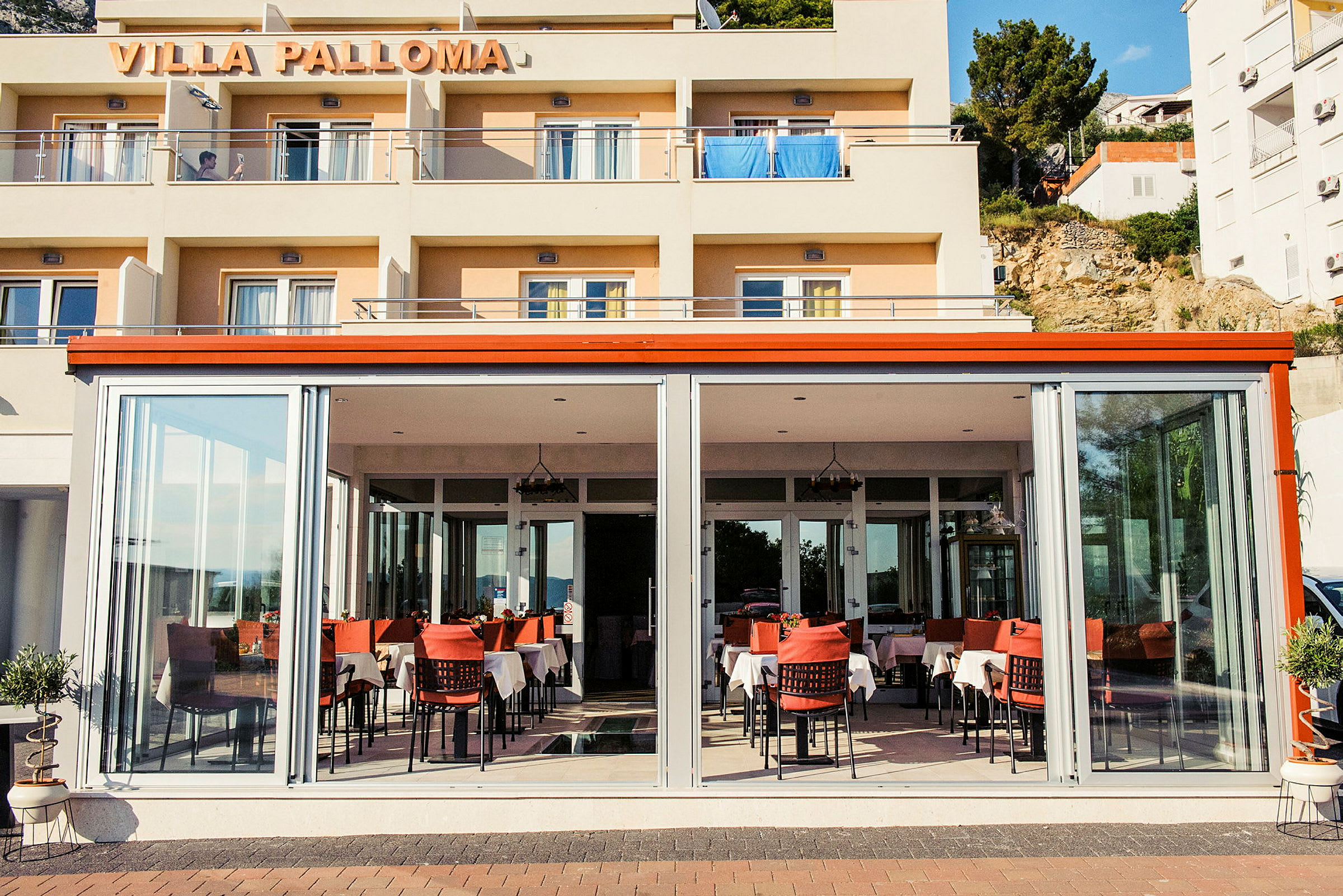 Indgangen til Villa Palloma med en restaurant fyldt med dækkede borde. (AI-genereret tekst)