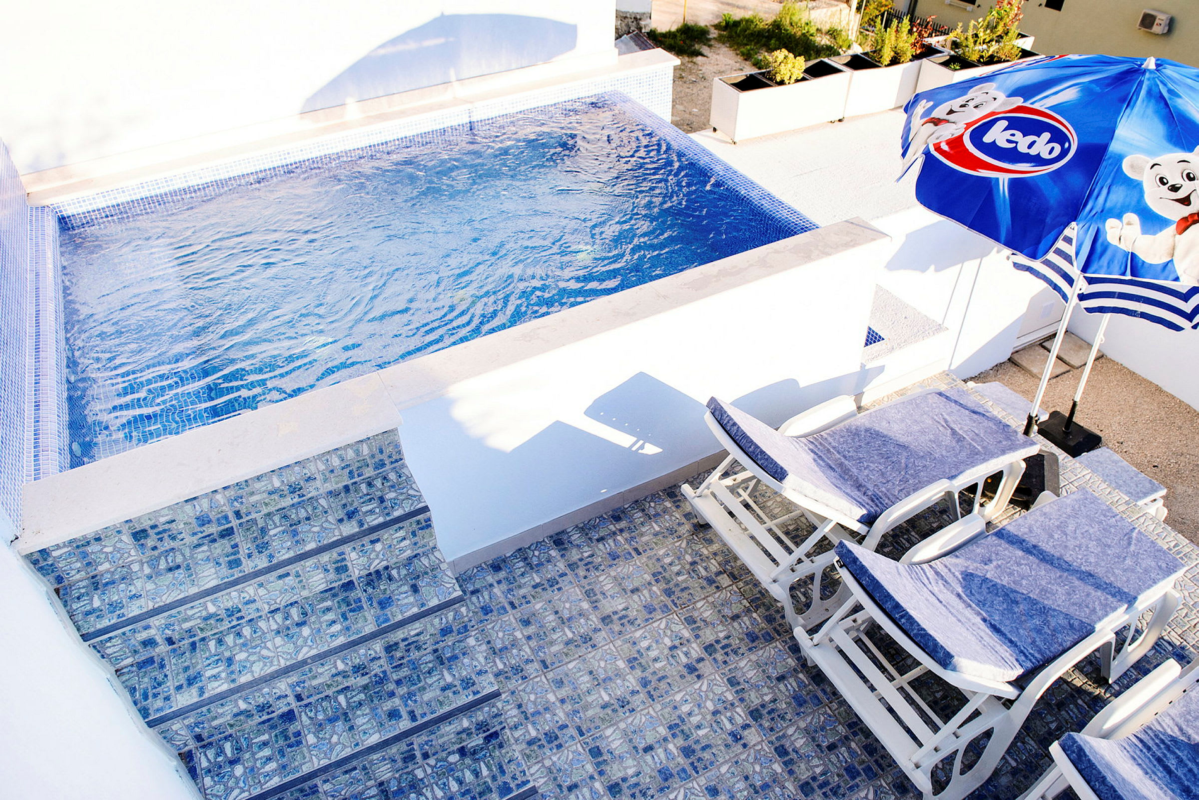Billede af en lille pool med blå mosaik, liggestole og en parasol. (AI-genereret tekst)