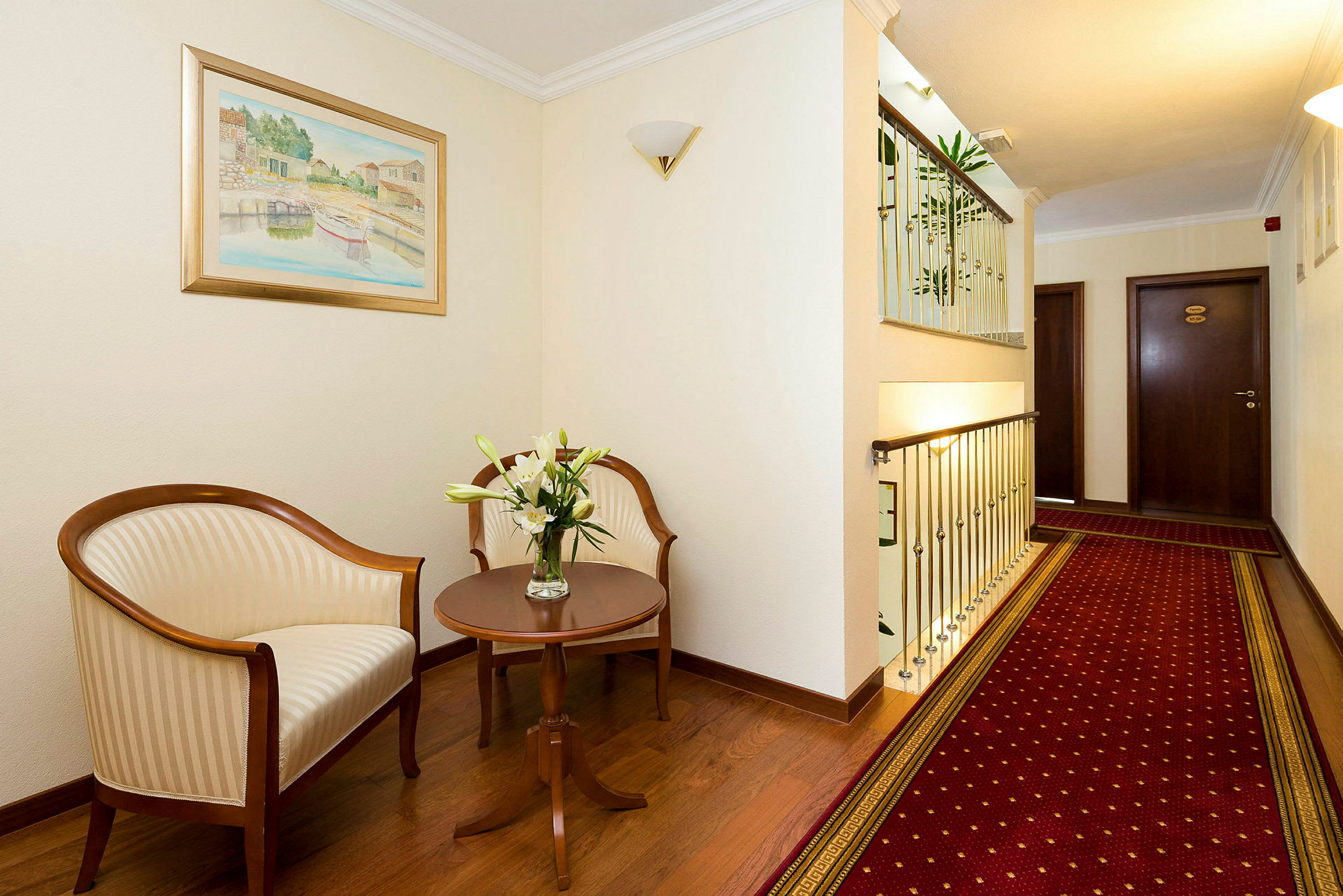 Billedet viser en hotelkorridor med siddeområde, inkluderer to stole, et sidebord med blomster og et rødt tæppe. (AI-genereret tekst)