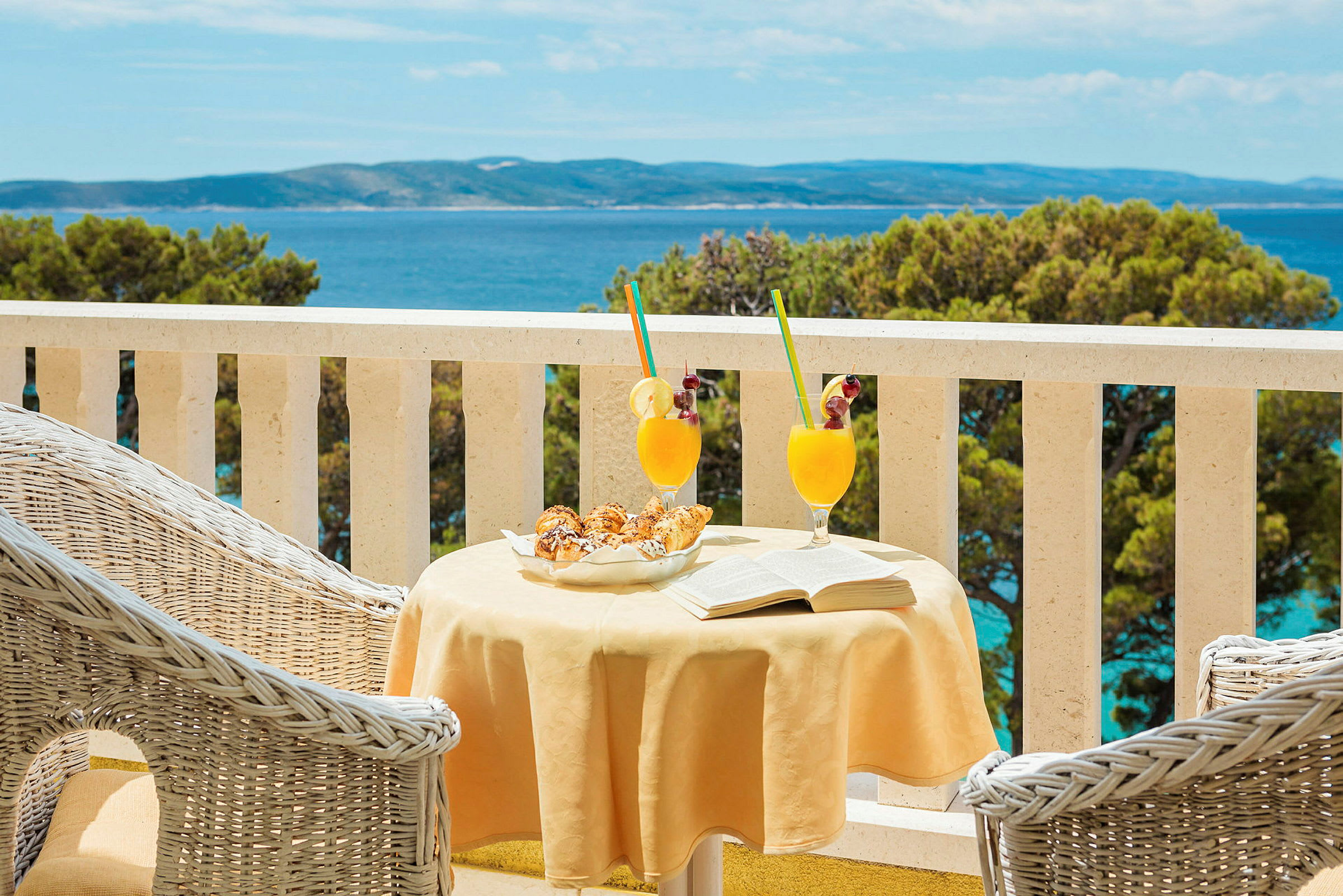 Et bord på en terrasse med to glas orange drik, bagværk og en åben bog. Havudsigt i baggrunden. (AI-genereret tekst)