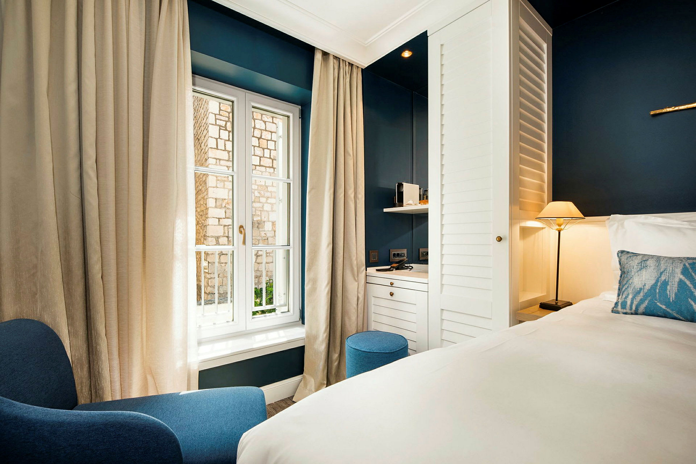 Et hotelværelse med moderne møbler, herunder en blå lænestol, en seng med hvidt sengetøj, en lille lampe og et skrivebord ved et vindue. (AI-genereret tekst)