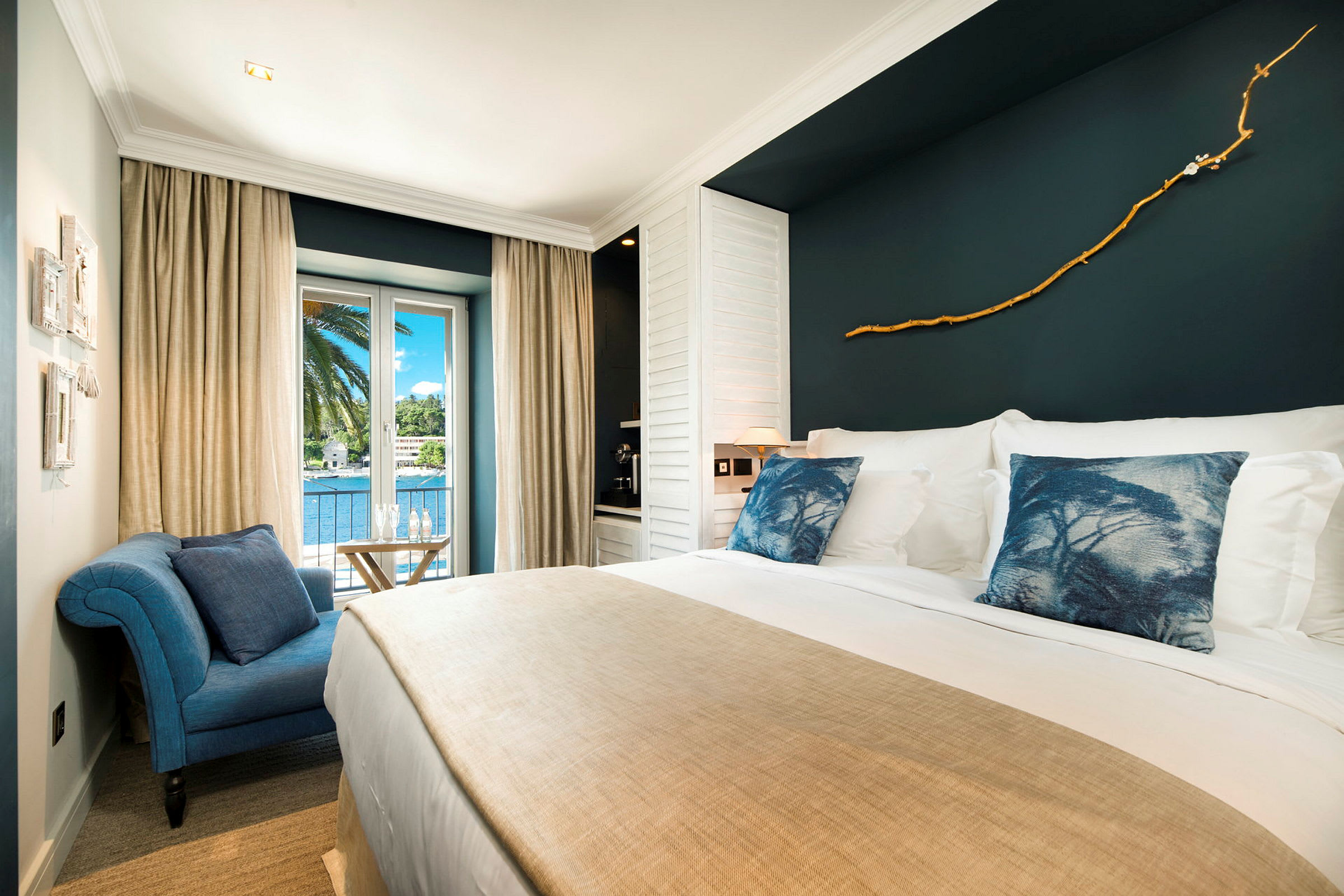 Et elegant hotelværelse med stor seng, blå pyntepuder, beige sengetæppe og dekoreret mørk væg. Værelset har et vindue med udsigt over palmer og vand. (AI-genereret tekst)