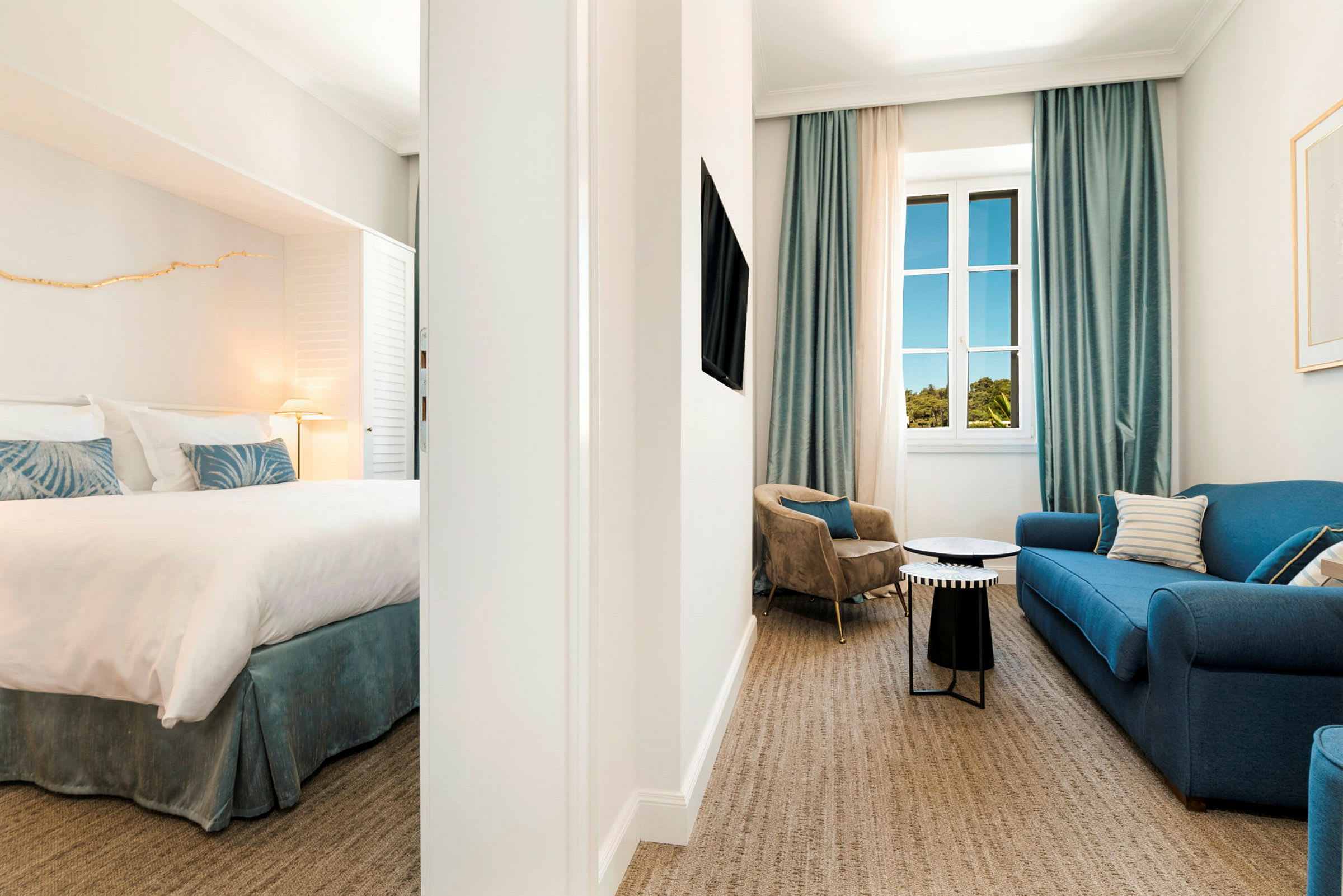 En hotel-suite med soveværelse og opholdsområde, indrettet med en blå sofa, lænestole og vindue med blå gardiner. (AI-genereret tekst)