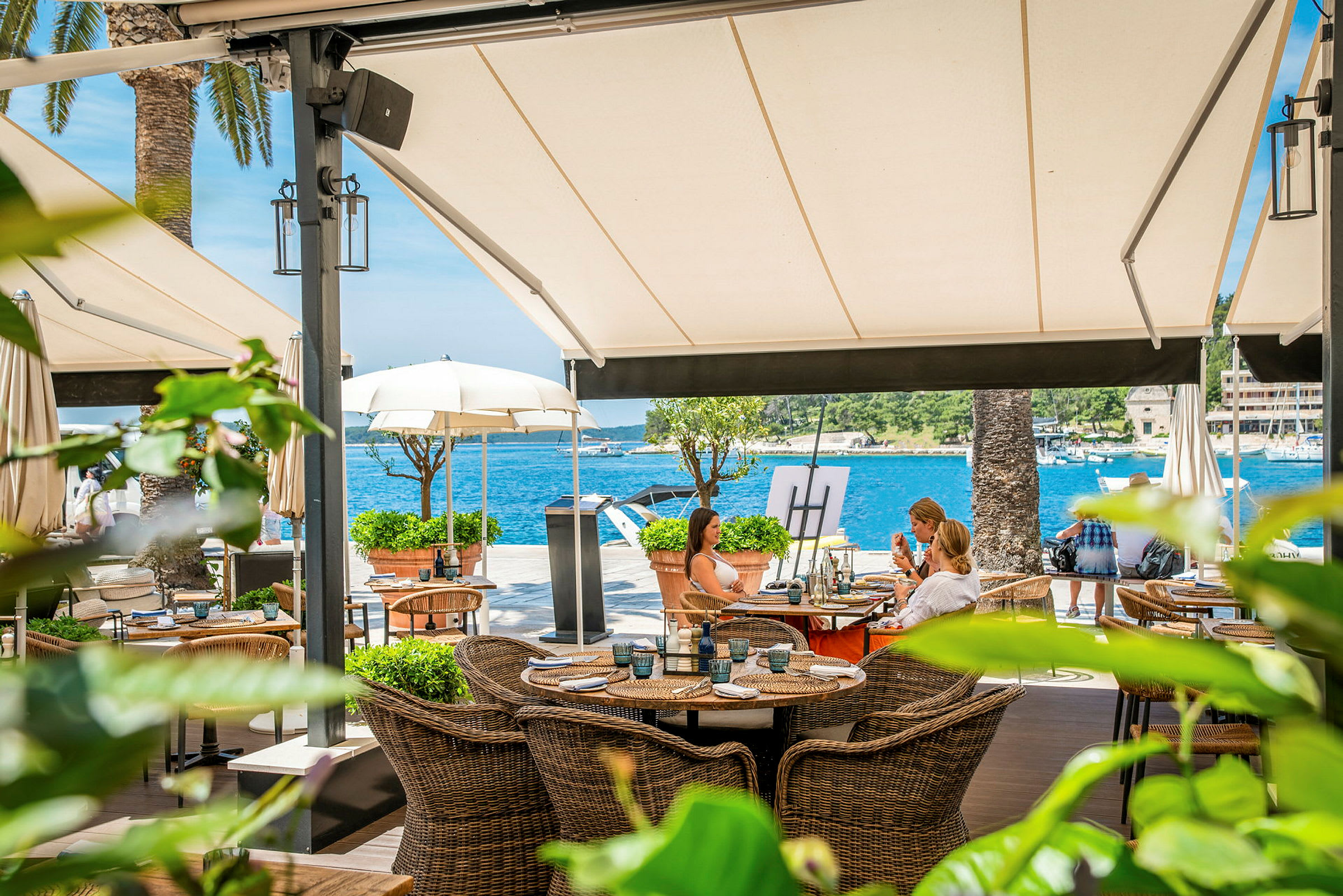 En restaurantterrasse ved vandet med kurvemøbler og udsigt over havet under store markiser. (AI-genereret tekst)