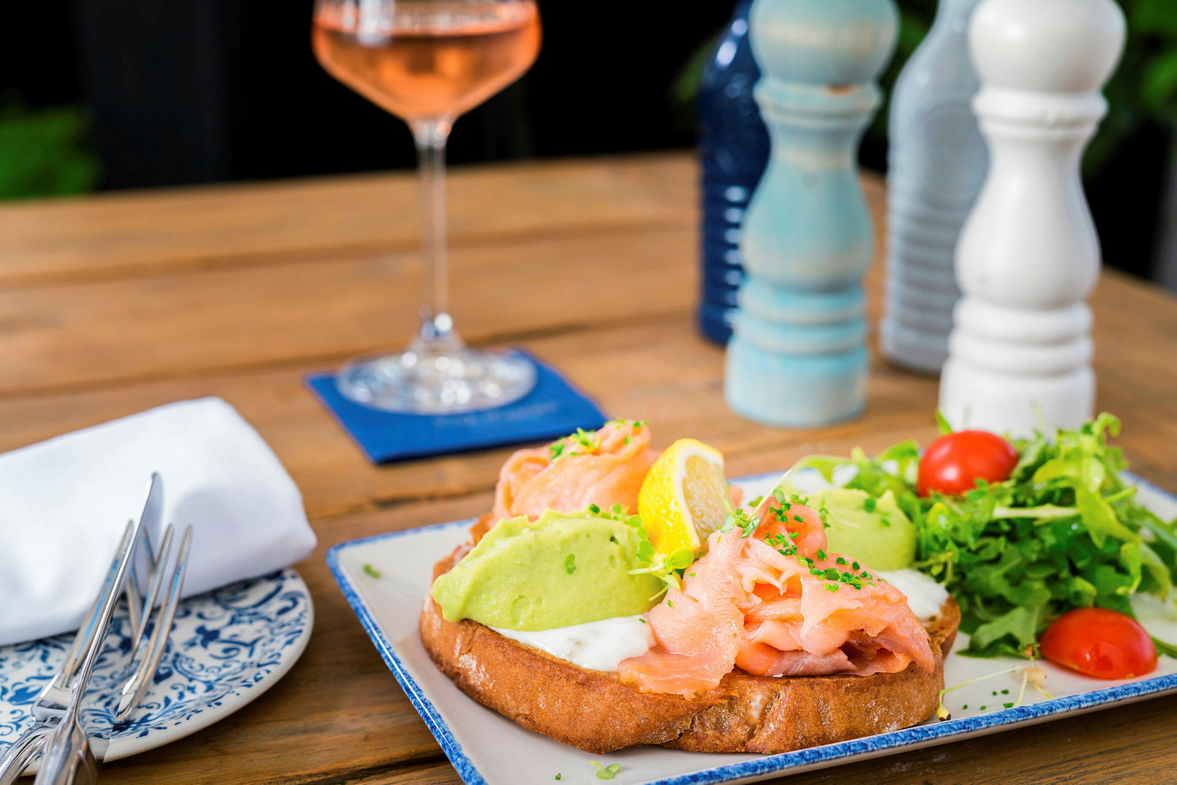 En tallerken med mad: smørrebrød med laks, avocado og grøntsager. I baggrunden er et vinglas og krydderkværne. (AI-genereret tekst)