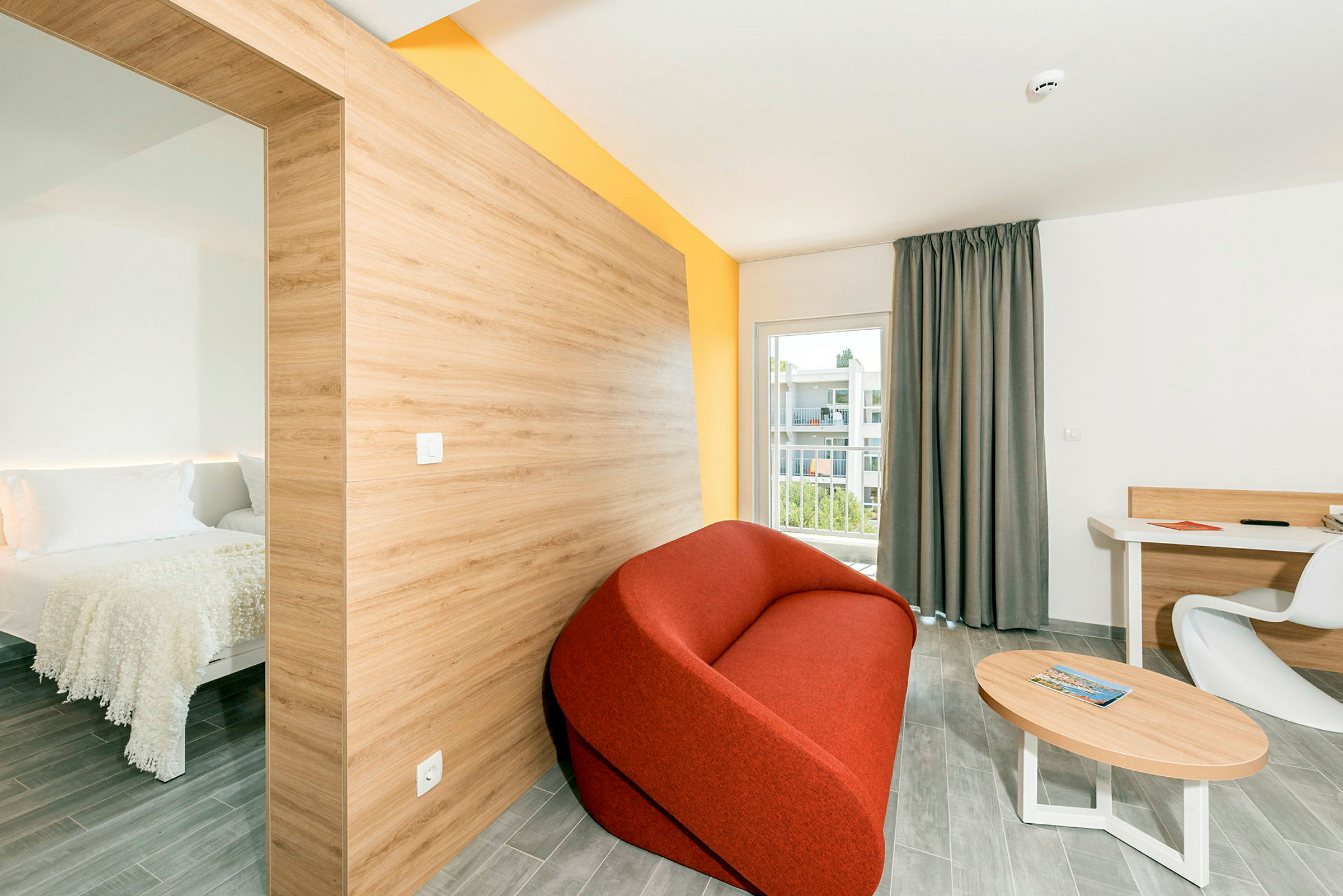 Et moderne hotelværelse med et separat soveområde til venstre, en rød sofa, et bord og en skriveplads med en stol samt en balkon med udsigt. (AI-genereret tekst)