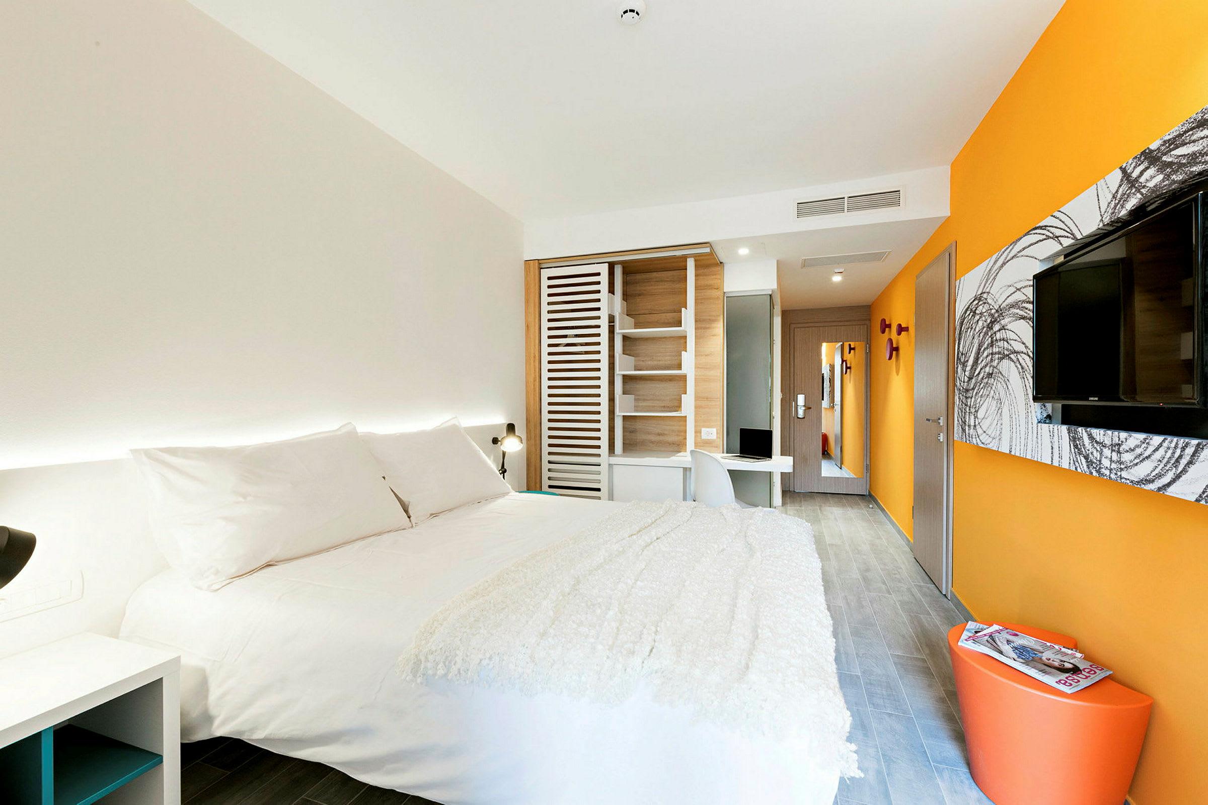 Et moderne hotelværelse med en stor seng med hvidt sengetøj, orange og hvide vægge, et vægmonteret TV og et skrivebord. (AI-genereret tekst)