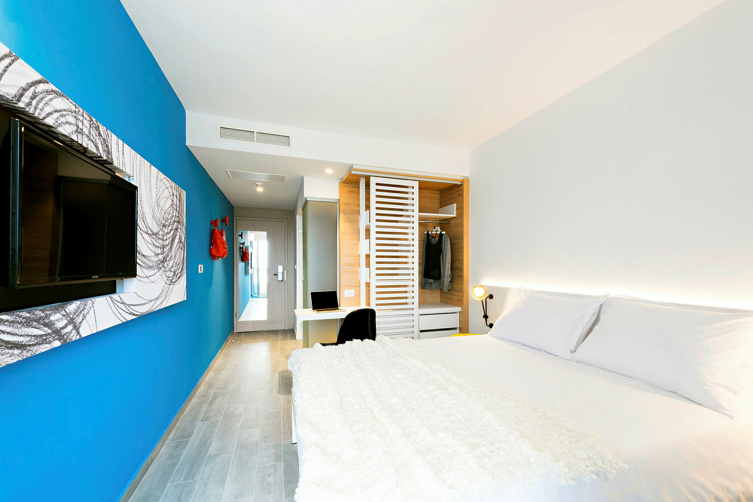 Et moderne hotelværelse med en stor seng, blå væg, tv, garderobe og et skrivebord. (AI-genereret tekst)