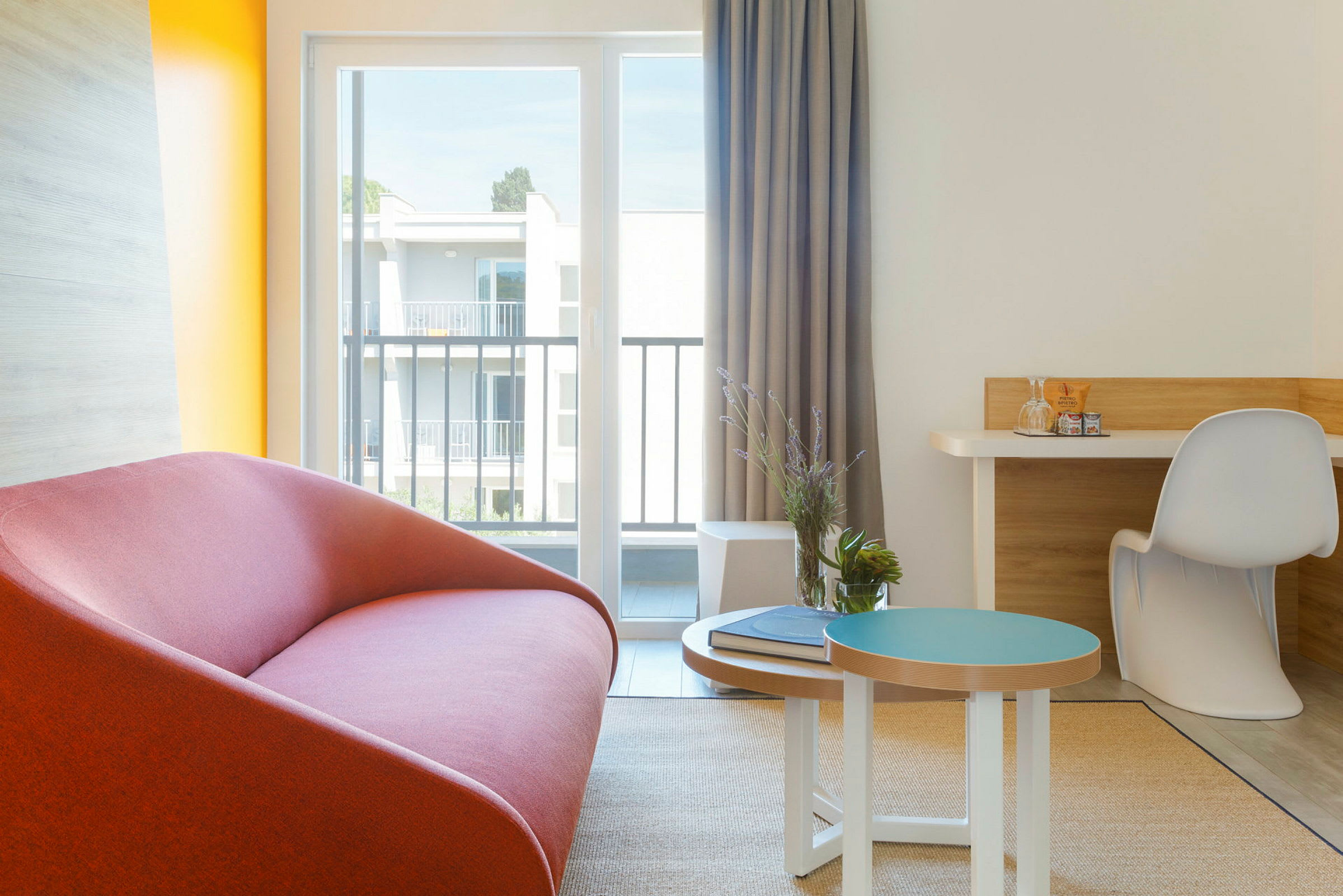 Et moderne indrettet hotelværelseshjørne med en rød sofa, et lille bord med dekorationer og en skrivebordskrog ved et stort vindue. (AI-genereret tekst)