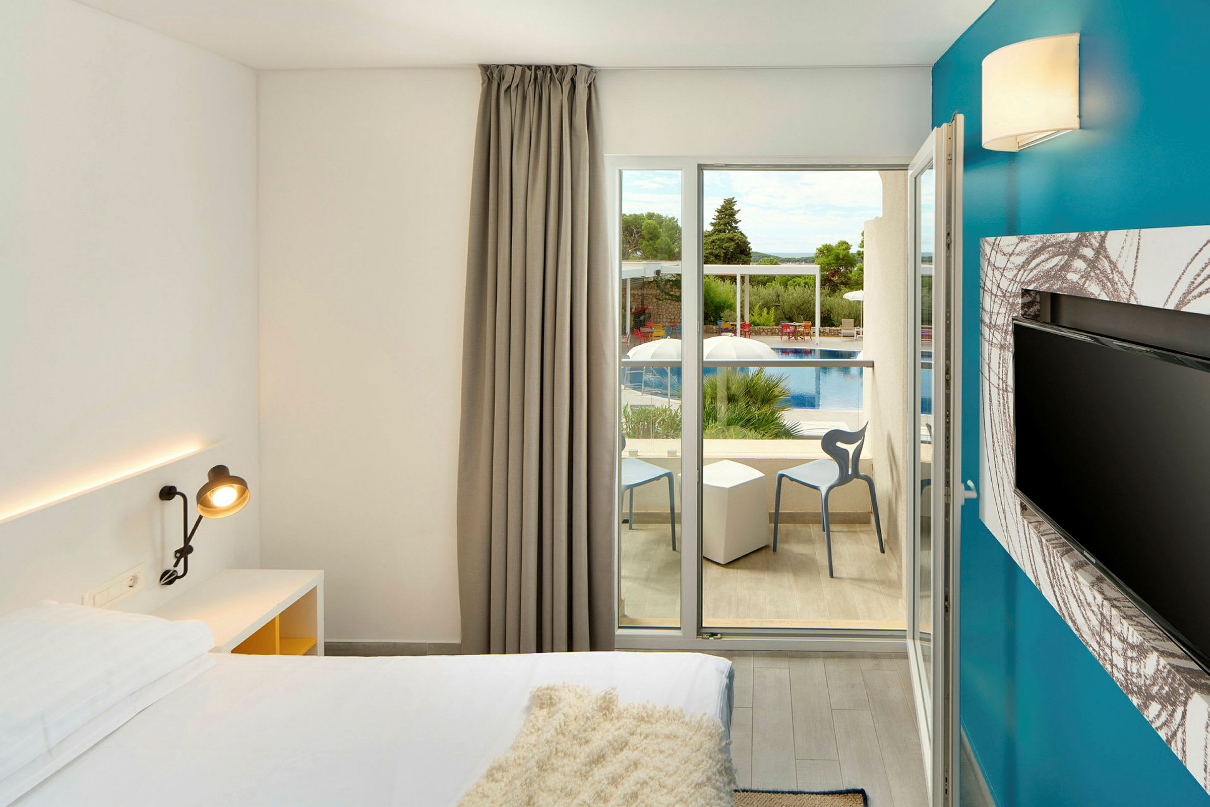 Et moderne hotelværelse med en seng, en skrivebordslampe og et fladskærms-TV. Vinduesdøre fører ud til en balkon med udsigt over en pool og grønne træer. (AI-genereret tekst)