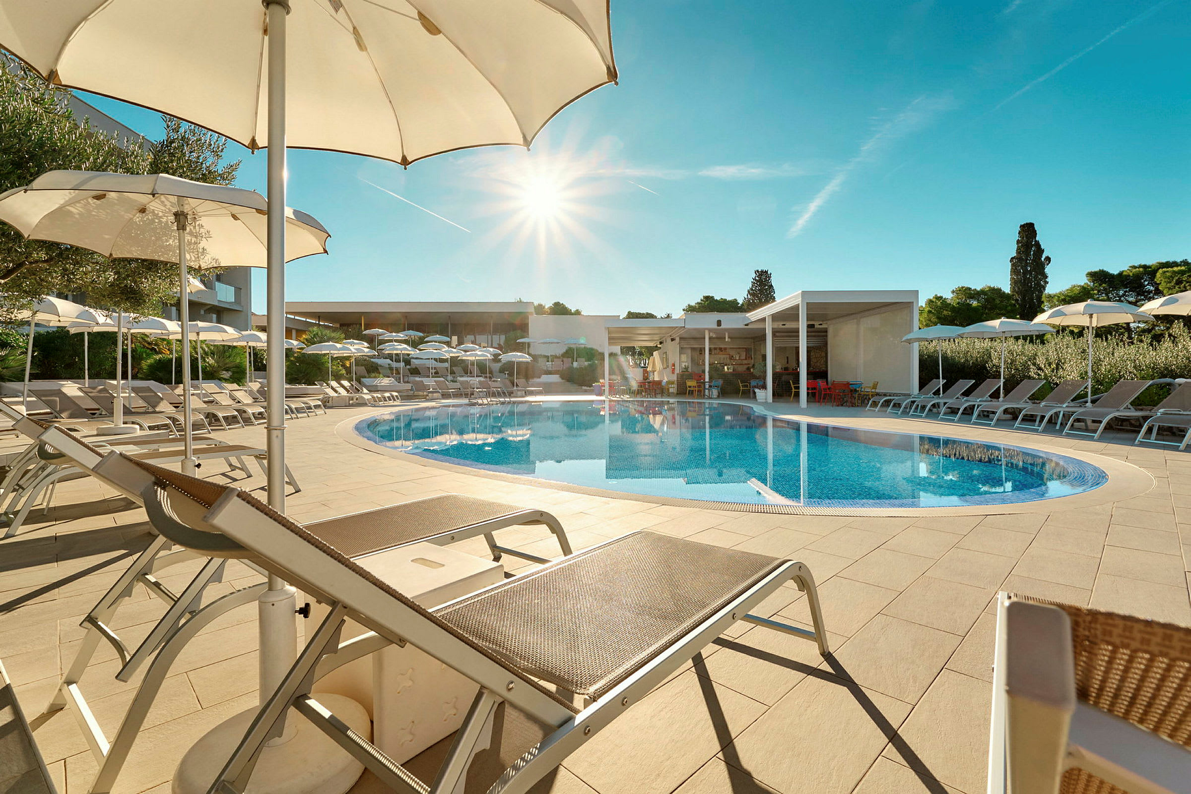 En udendørs swimmingpool omgivet af liggestole og parasoller under en klar blå himmel. (AI-genereret tekst)
