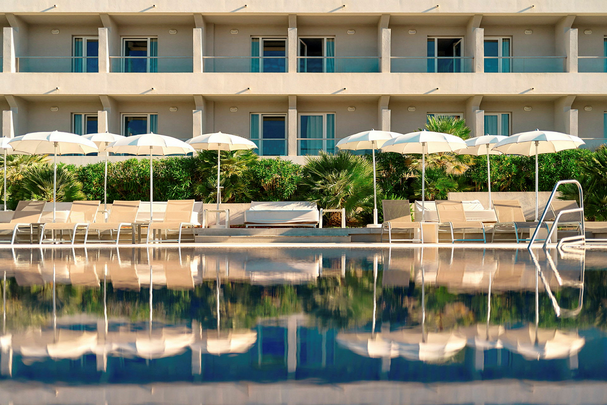 Billede af en oplyst hotelfacade med altaner bag en pool. Foran poolen er der solsenge og hvide parasoller samt bevoksning. (AI-genereret tekst)