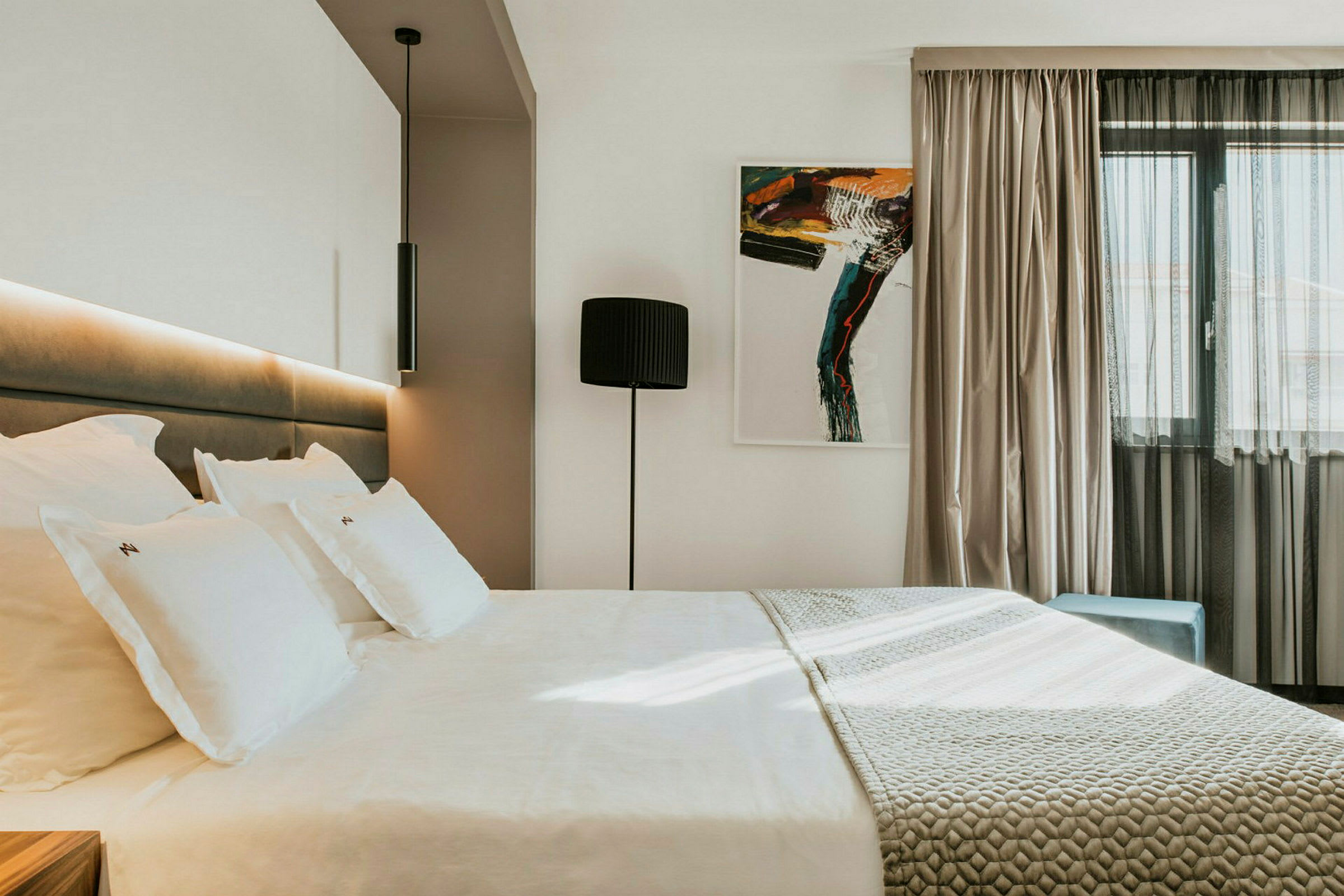 Moderne hotelværelse med en stor seng, kunstværk på væggen og et vindue med lyse gardiner. (AI-genereret tekst)