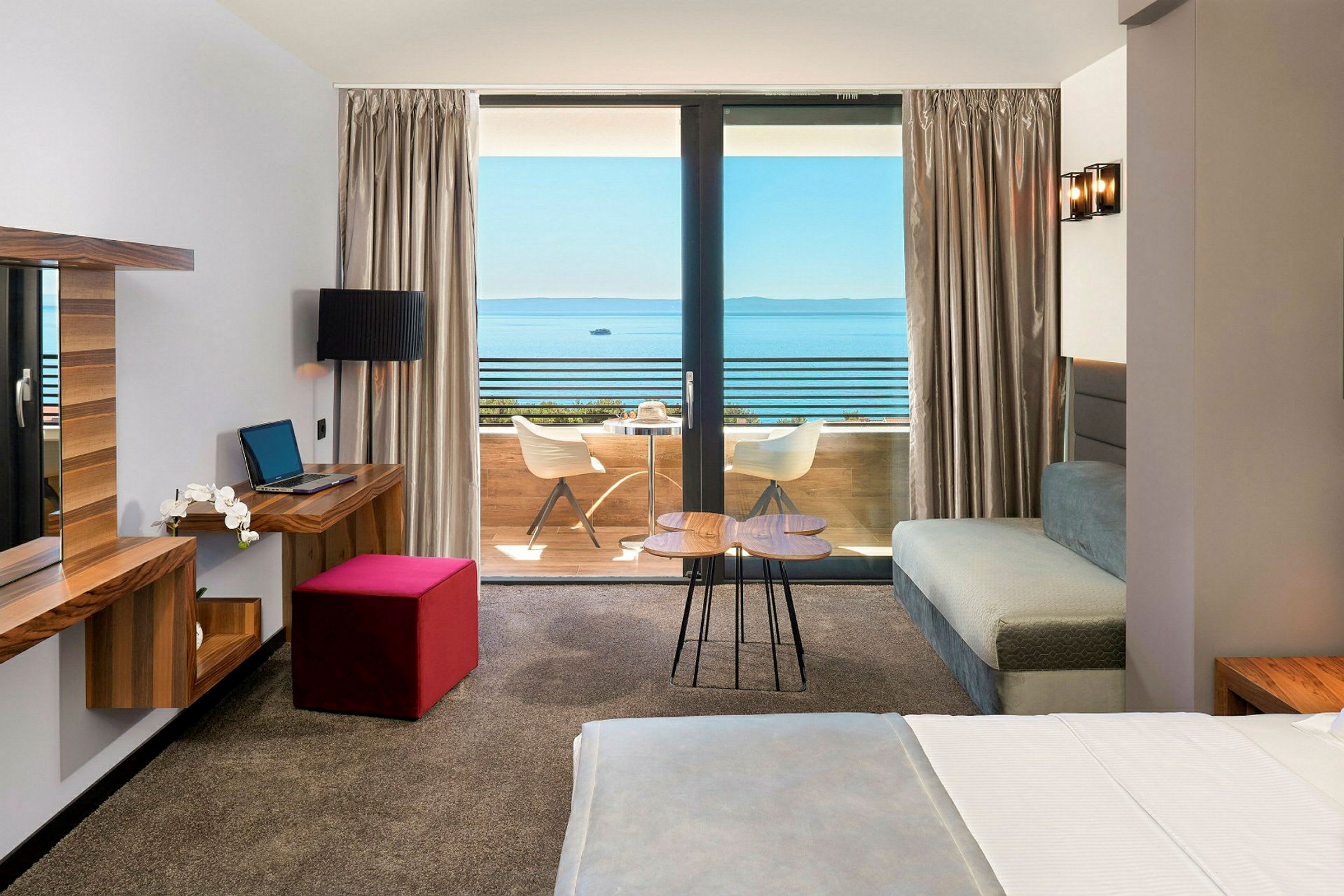 Hotelværelse med seng, skrivebord og udsigt til en altan og havet. (AI-genereret tekst)