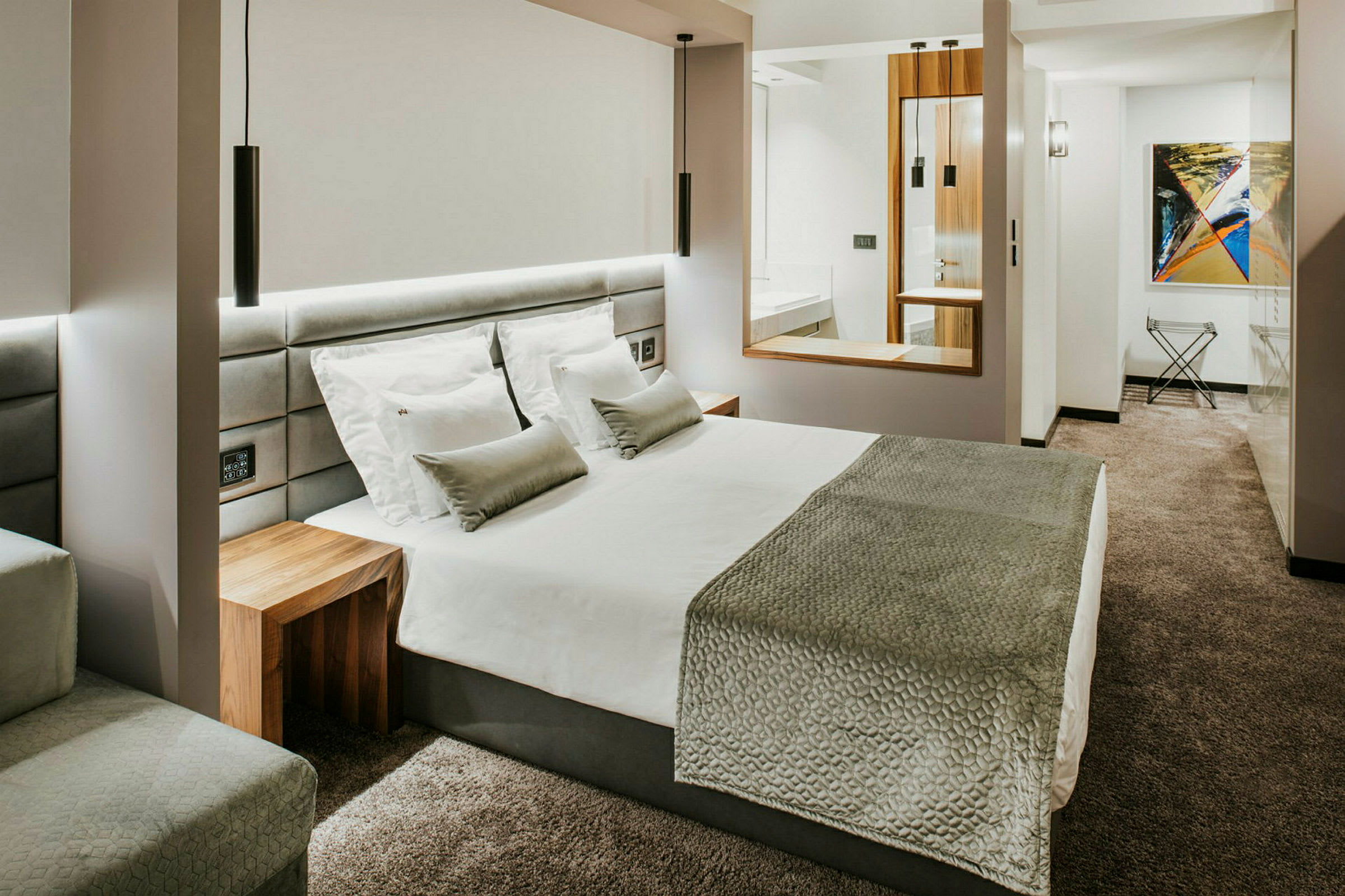 Et moderne hotelværelse med en stor seng, hvide sengetøj, gråt sengetæppe og puder samt gråt væg-til-væg tæppe. (AI-genereret tekst)