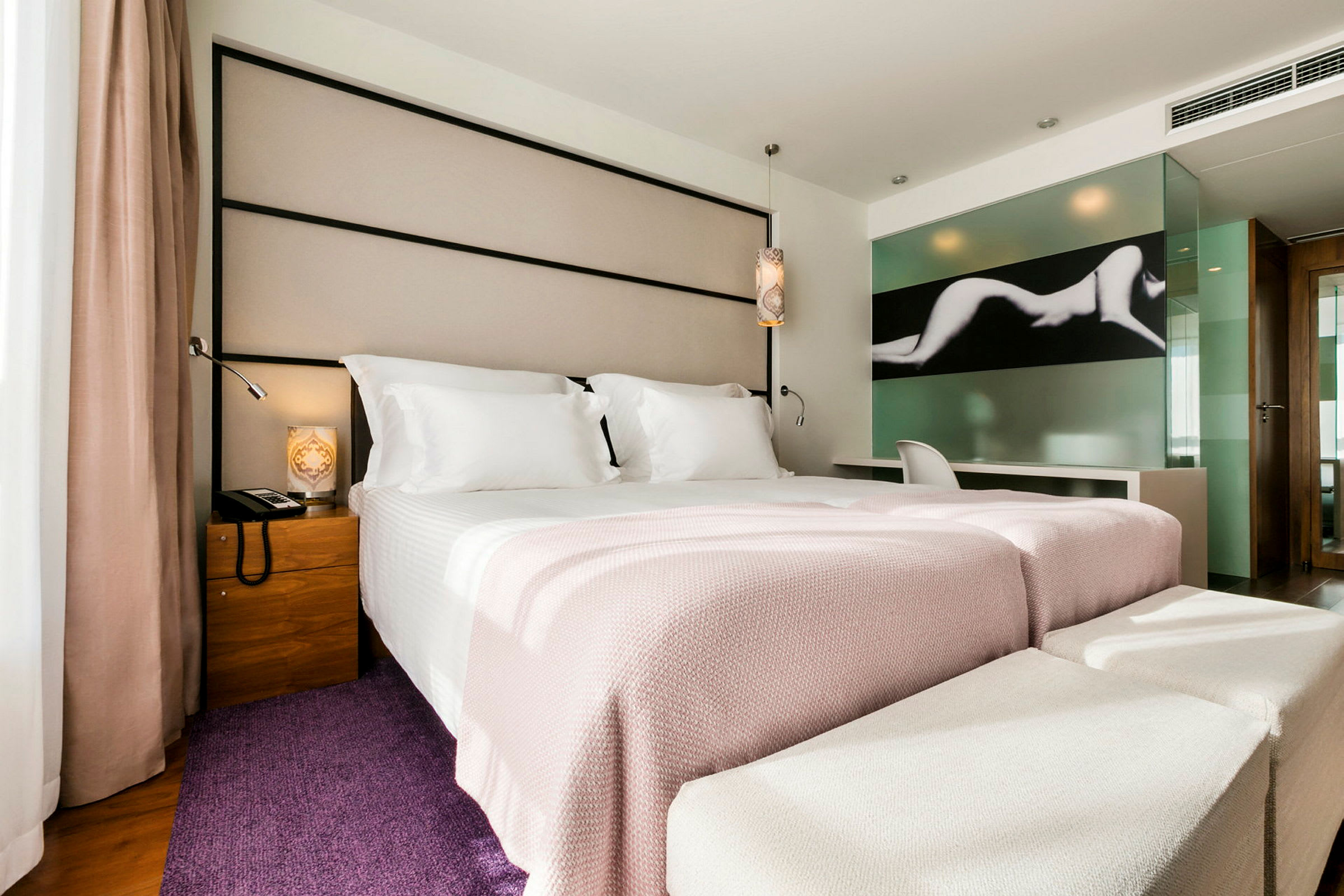 Et moderne hotelværelse med en bred seng redt med hvidt og lyserødt sengetøj. Der er en stol og et skrivebord ved en glasvæg med kunstmotiv. (AI-genereret tekst)