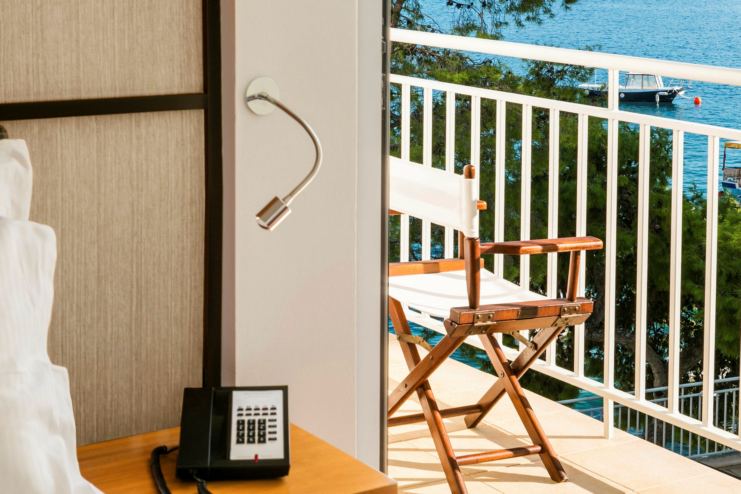 En del af et hotelværelse med en bord med en telefon og en læselampe, samt en balkon med udsigt til havet og en stol. (AI-genereret tekst)