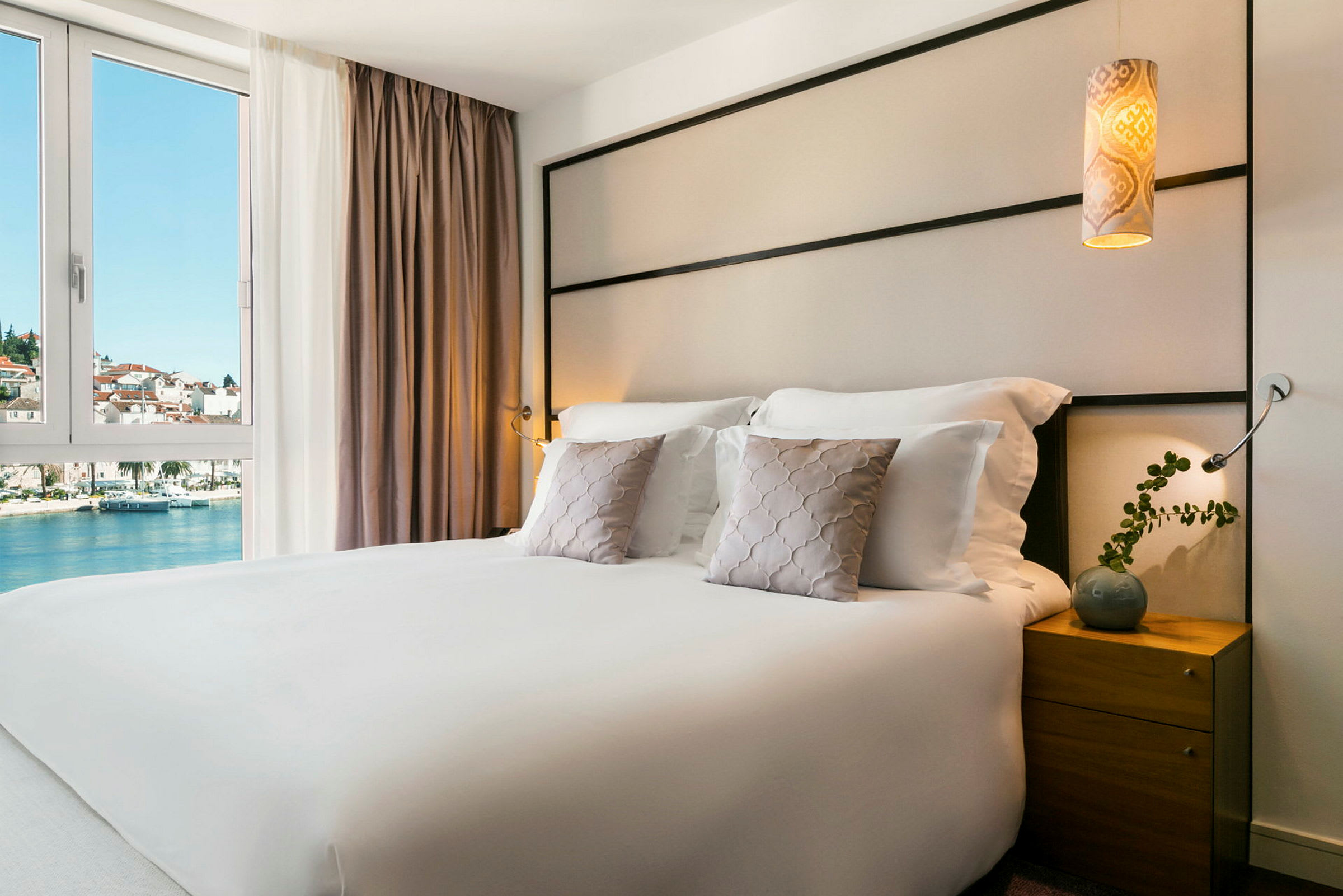 Hotelværelse med en dobbeltseng, pyntepuder og et natbord. Udsigt til vand og bygninger gennem vinduet. (AI-genereret tekst)