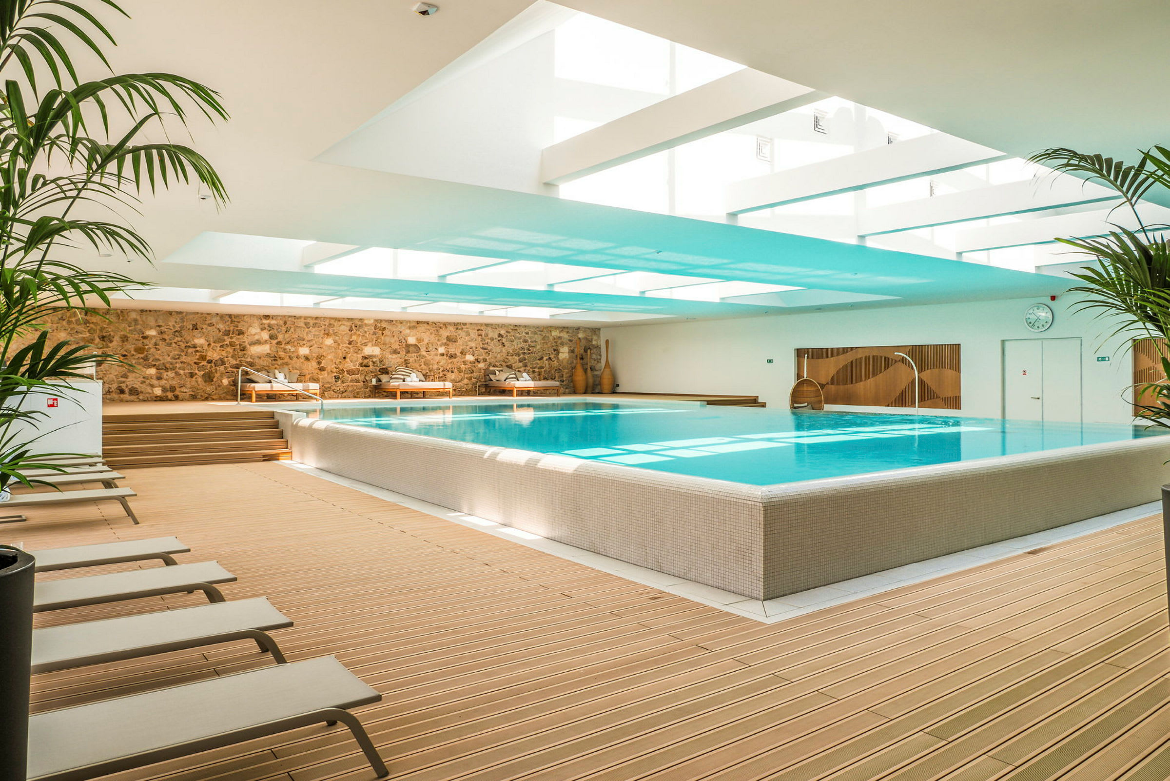 En indendørs swimmingpool omgivet af liggestole og planter. (AI-genereret tekst)