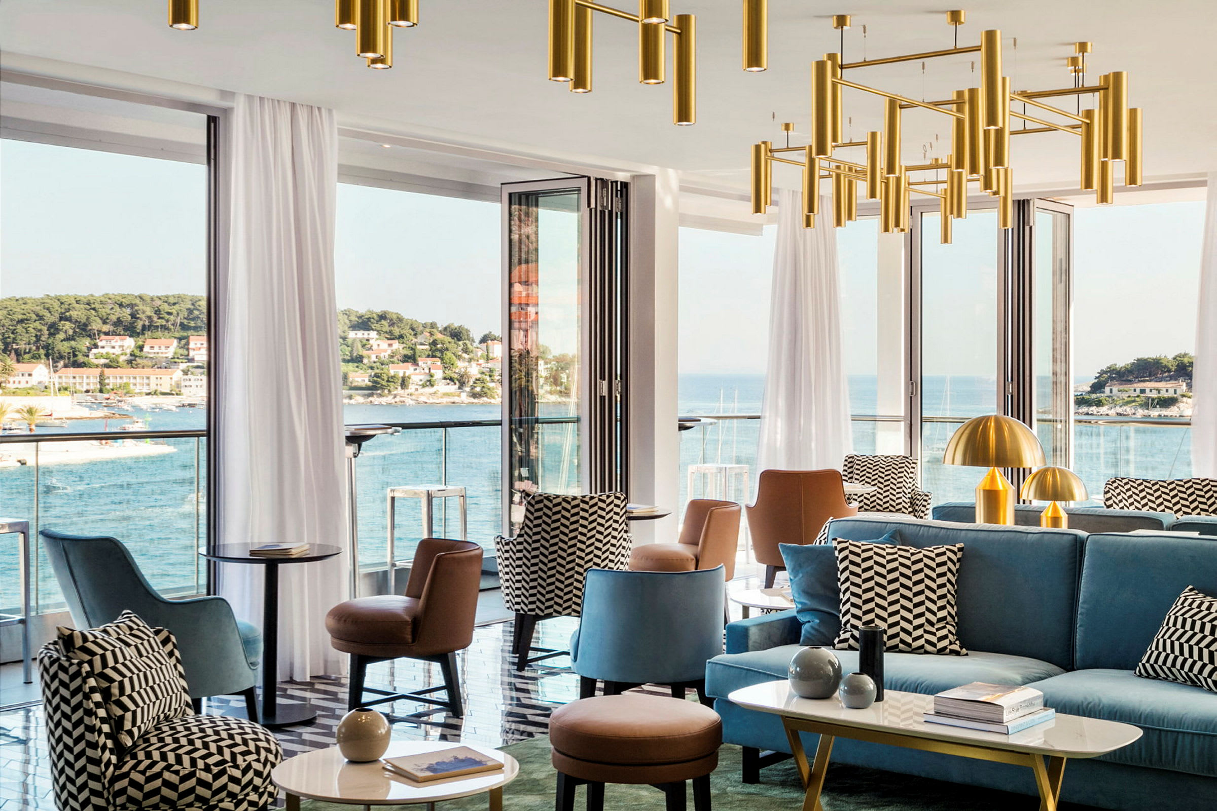 En moderne lounge med siddepladser og dekorative lysekroner, omgivet af store vinduer med udsigt over en kystby og havet. (AI-genereret tekst)