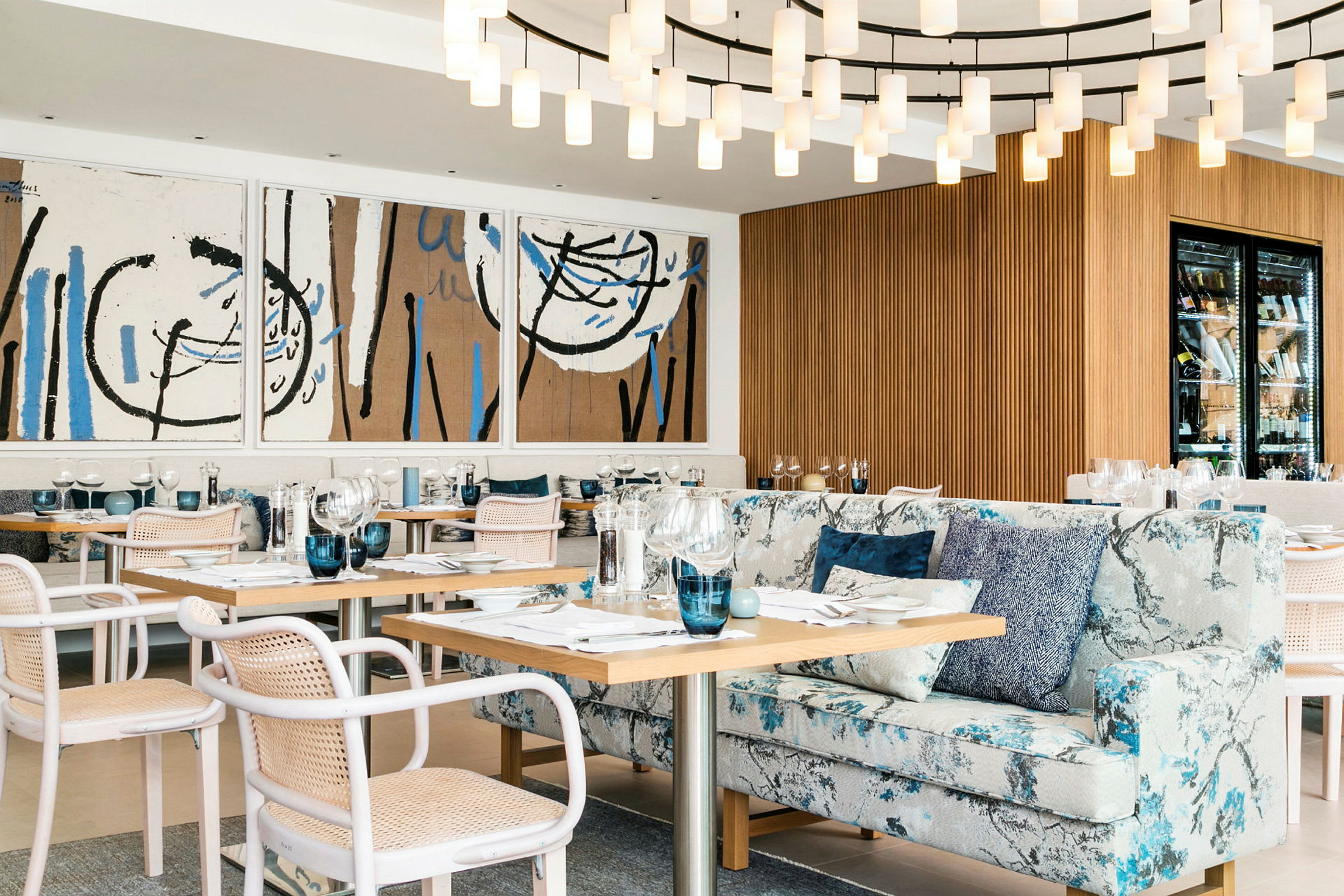 Et elegant indrettet restaurantområde med moderne kunstværker på væggen, en stor lysekrone, komfortable stole og sofaer samt opdækkede borde. (AI-genereret tekst)