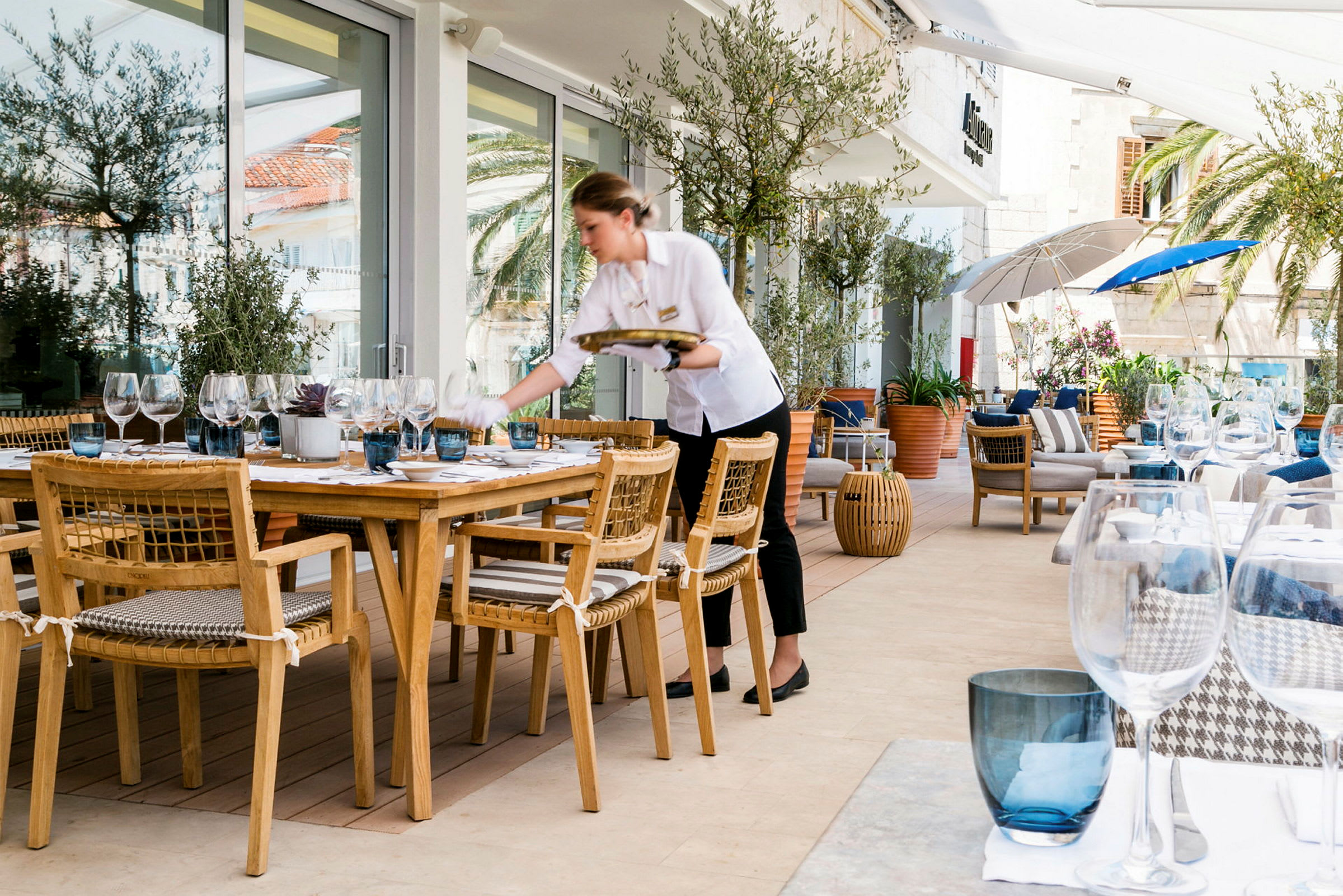En udendørs restaurant med træborde og stole samt opdækning med glas og porcelæn. Smuk omgivelser med planter og parasoller. (AI-genereret tekst)