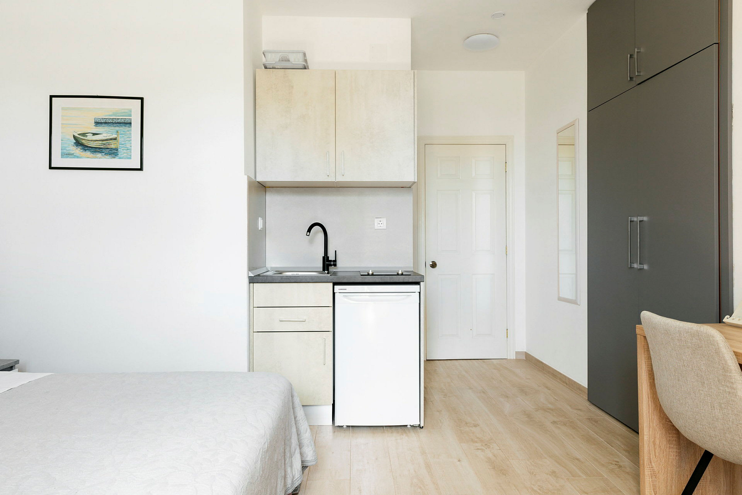 Velindrettet værelse med minimalistisk interiør inklusive en seng, tekøkken, garderobe og skrivebordsstol. (AI-genereret tekst)