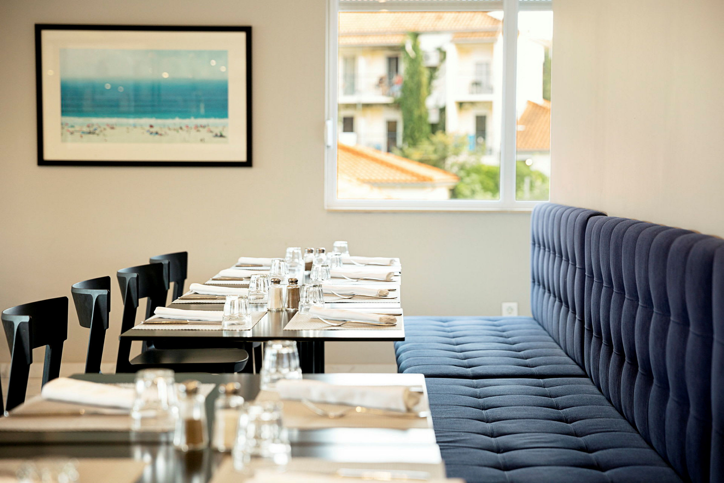 Et restaurantmiljø med dækkede borde med glas, bestik og servietter. En blå polstret sofa og vindue, der viser bygninger udenfor. Et billede med strandmotiv på væggen. (AI-genereret tekst)