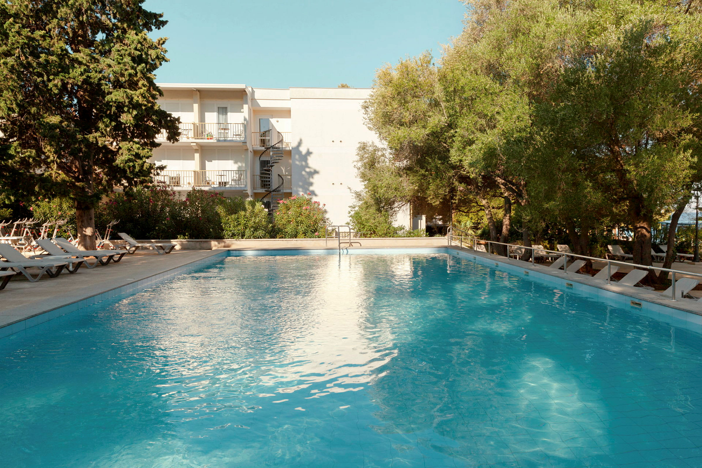 Udendørs pool ved et hotel omgivet af liggestole og grønne områder. (AI-genereret tekst)