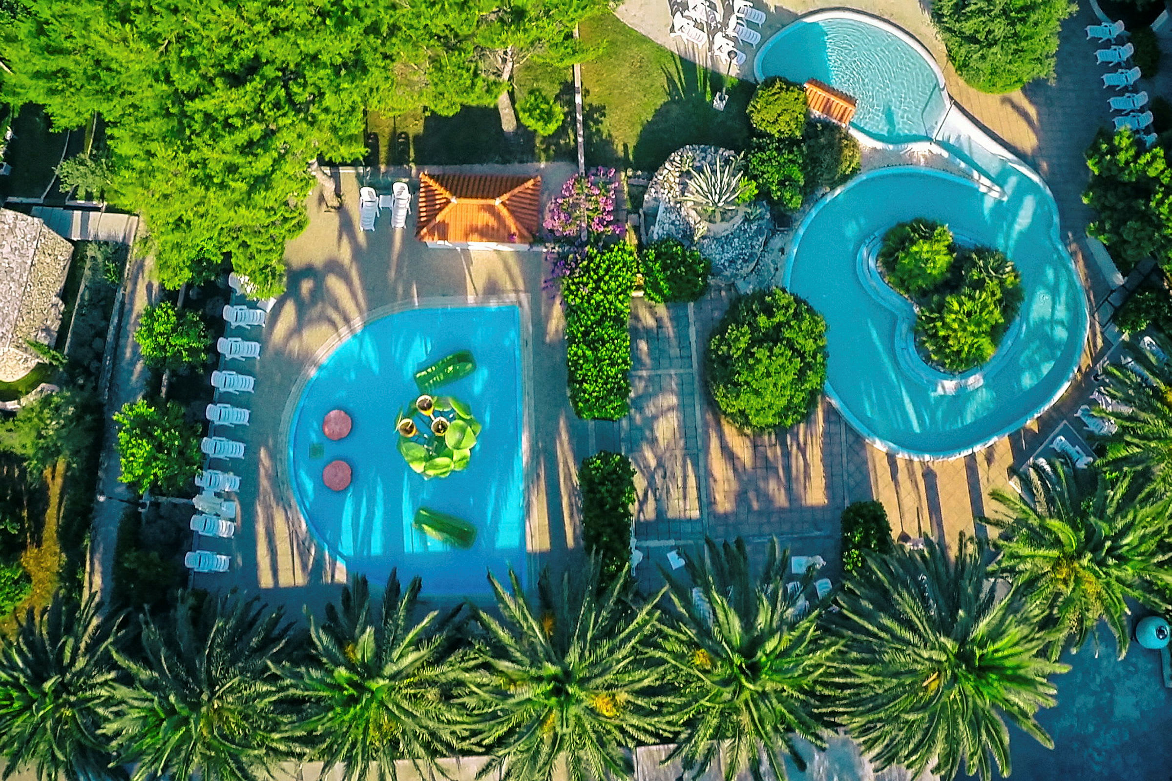 Udsigt oppefra over to swimmingpools omgivet af stole, vegetation og gangstier. (AI-genereret tekst)