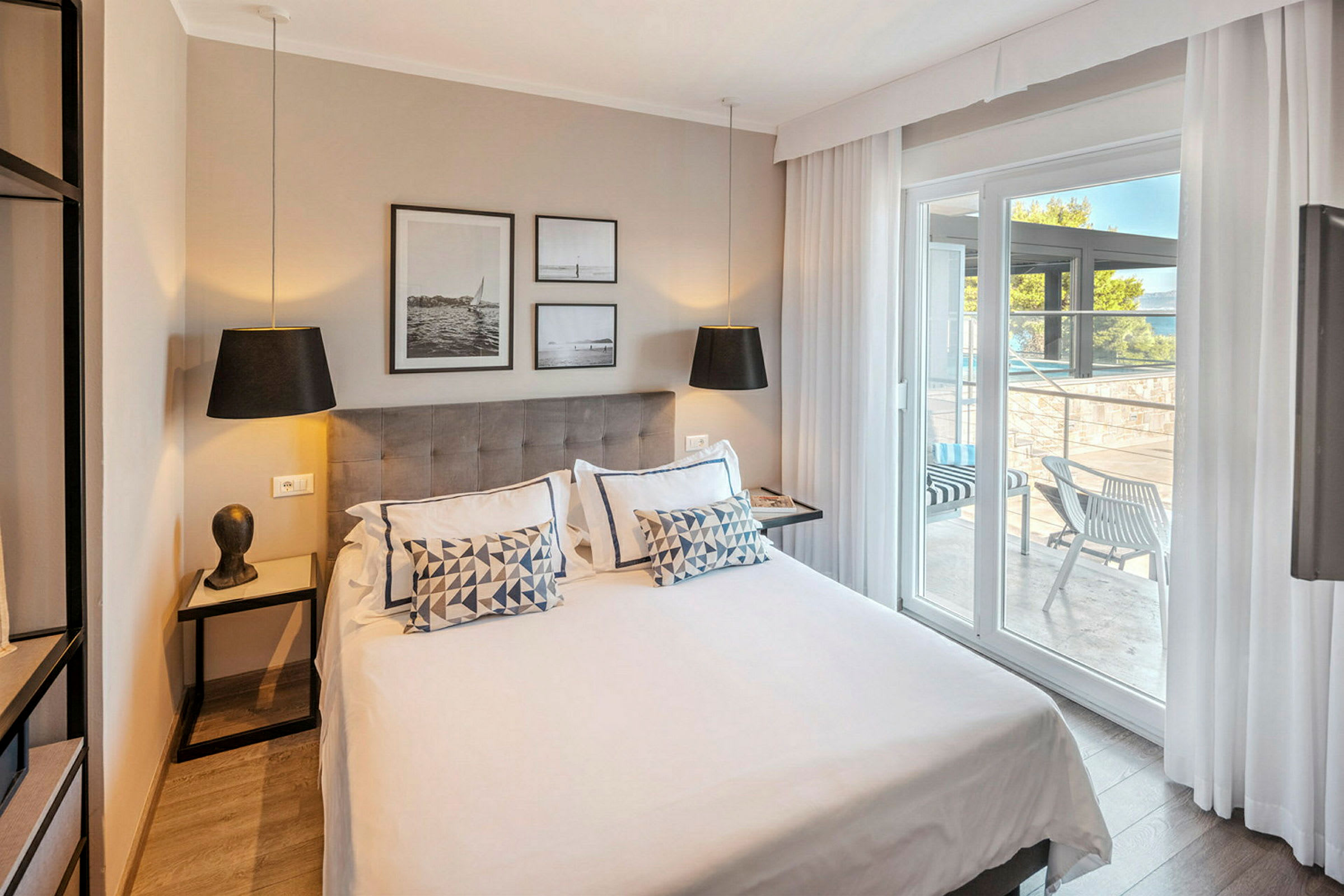 Moderne hotelværelse med dobbeltseng, dekoreret med puder og kunst på væggen. Glasdøre fører ud til en møbleret terrasse. (AI-genereret tekst)