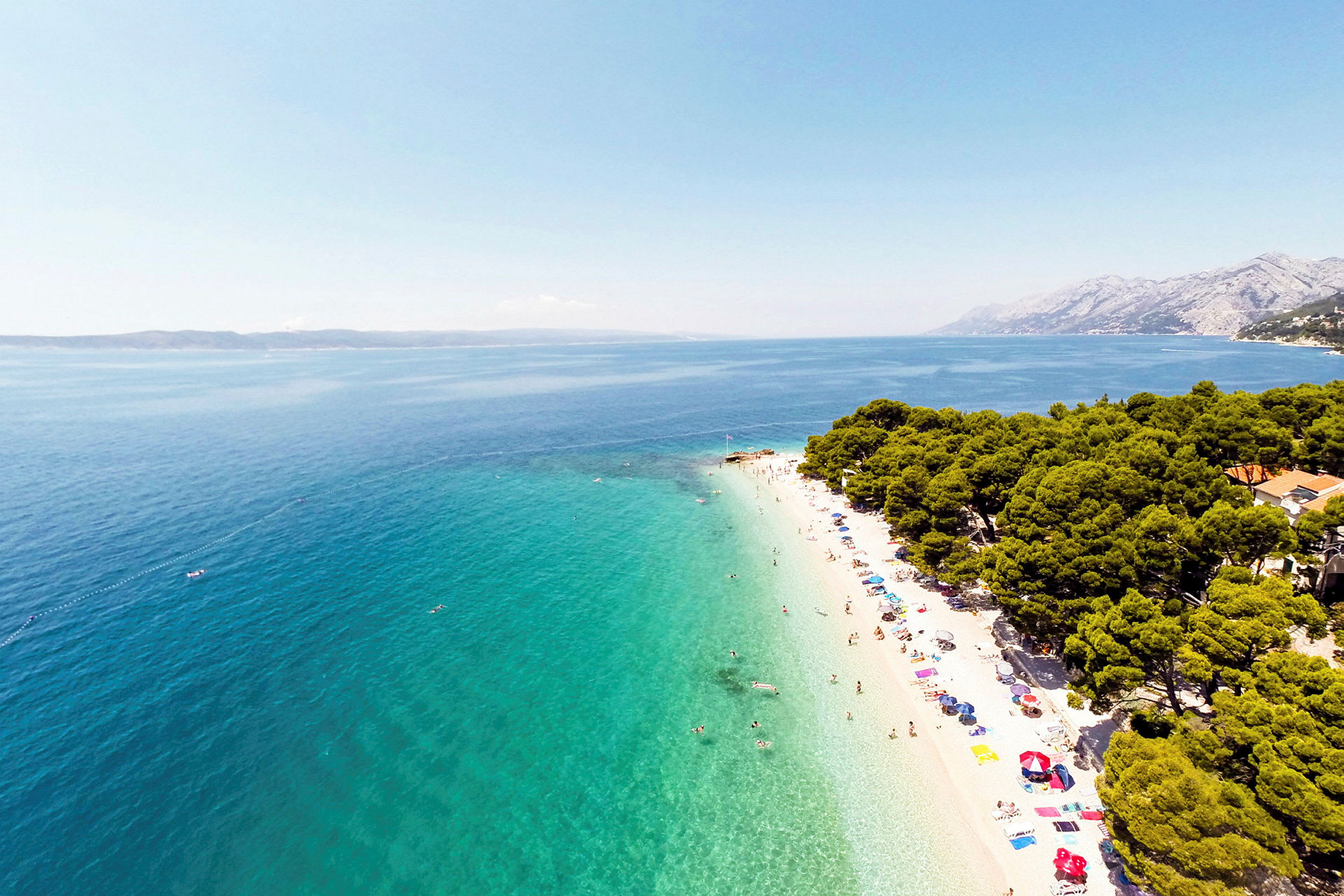 Strand med turkis vand, parasoller og badende gæster, omgivet af grønne træer. (AI-genereret tekst)