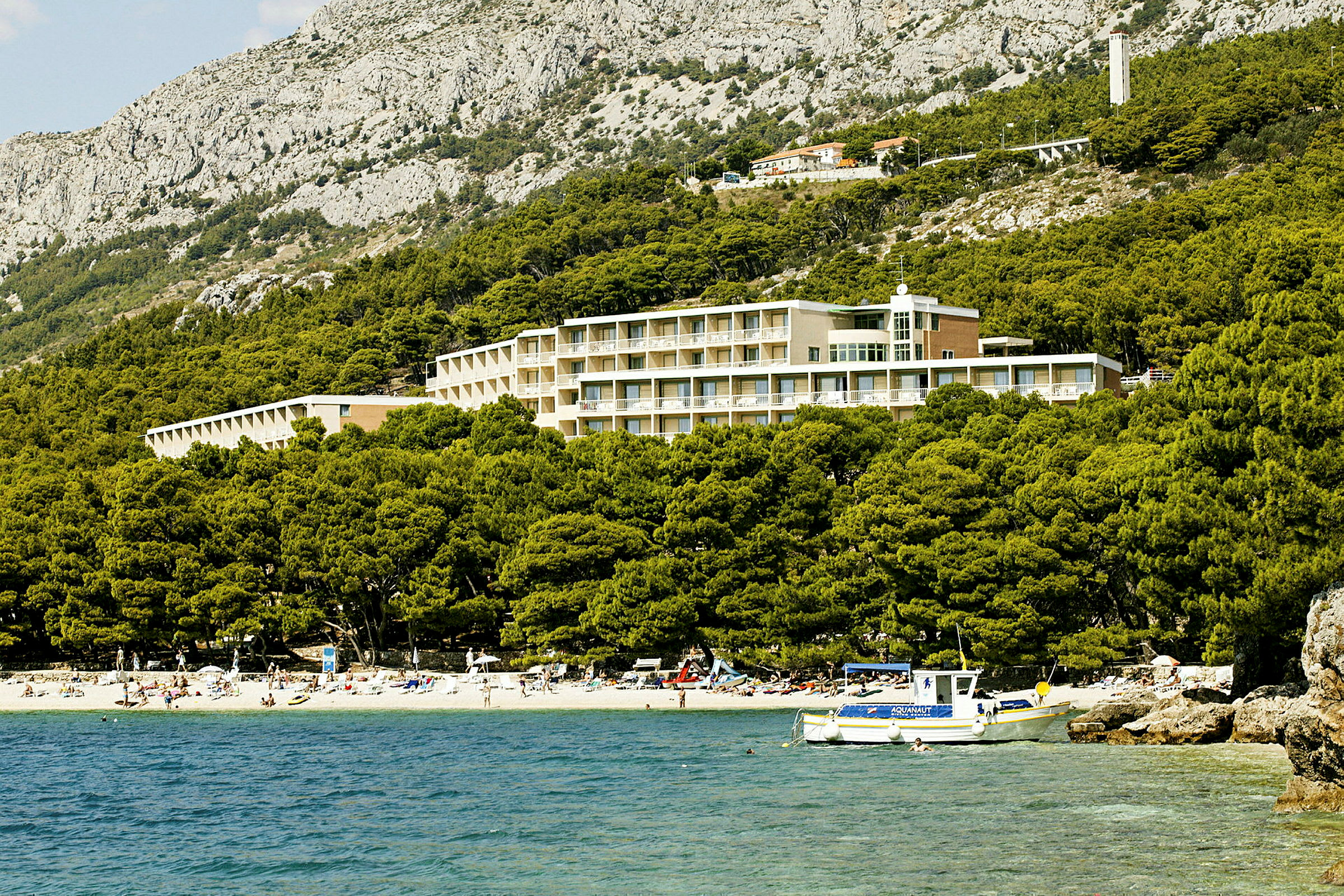 Hotel beliggende ved en fyrreskov og strand i et bjergrigt landskab. (AI-genereret tekst)