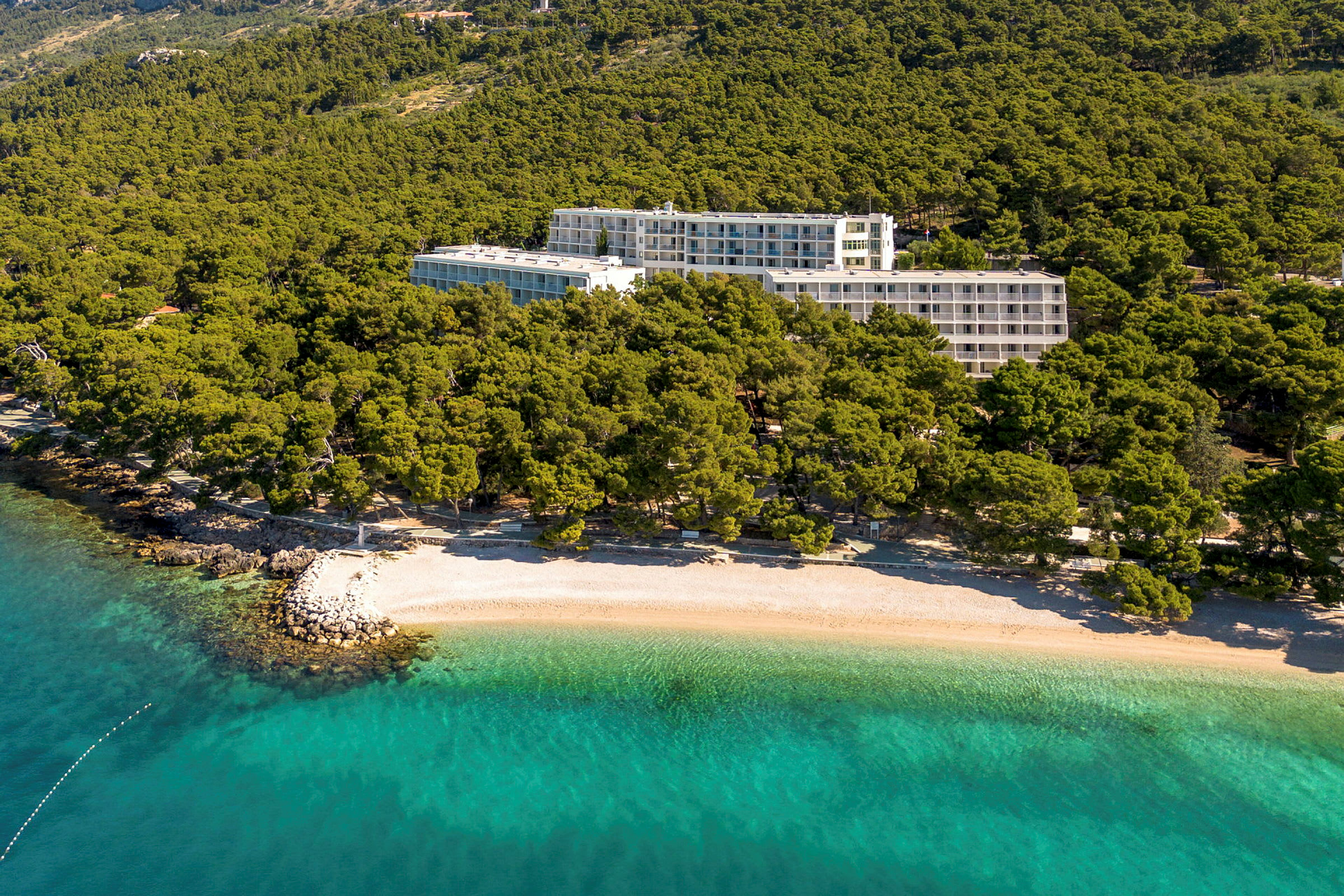 Hotelbygninger omgivet af grønt nær en strand med klart turkis vand. (AI-genereret tekst)