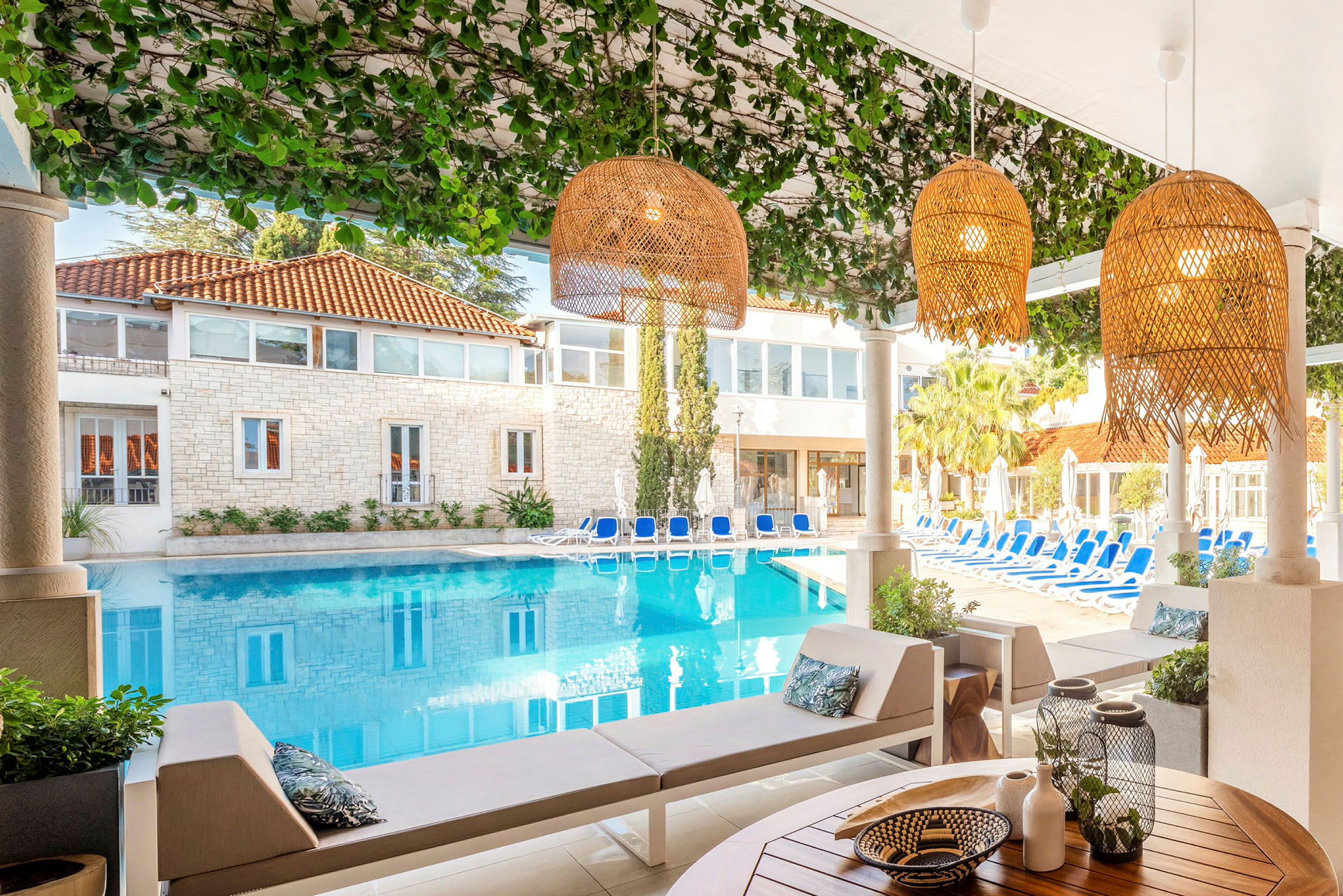 Udsigt over en pool omgivet af solsenge og et hus med stenfacade, fotograferet fra et skyggefuldt område med siddepladser og dekorationer. (AI-genereret tekst)