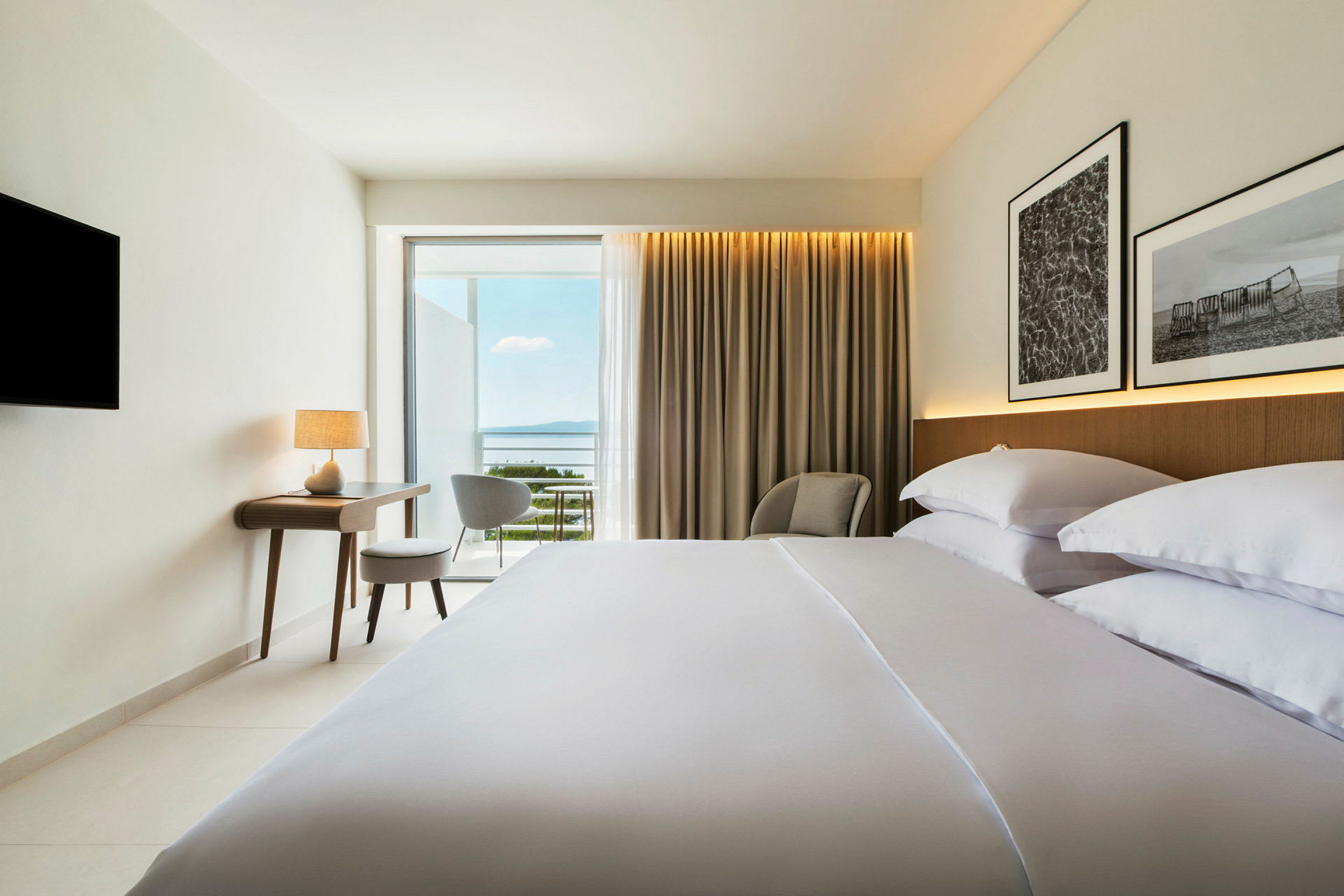 Hotelværelse med redt seng, skrivebord, stol og udsigt over havet. (AI-genereret tekst)