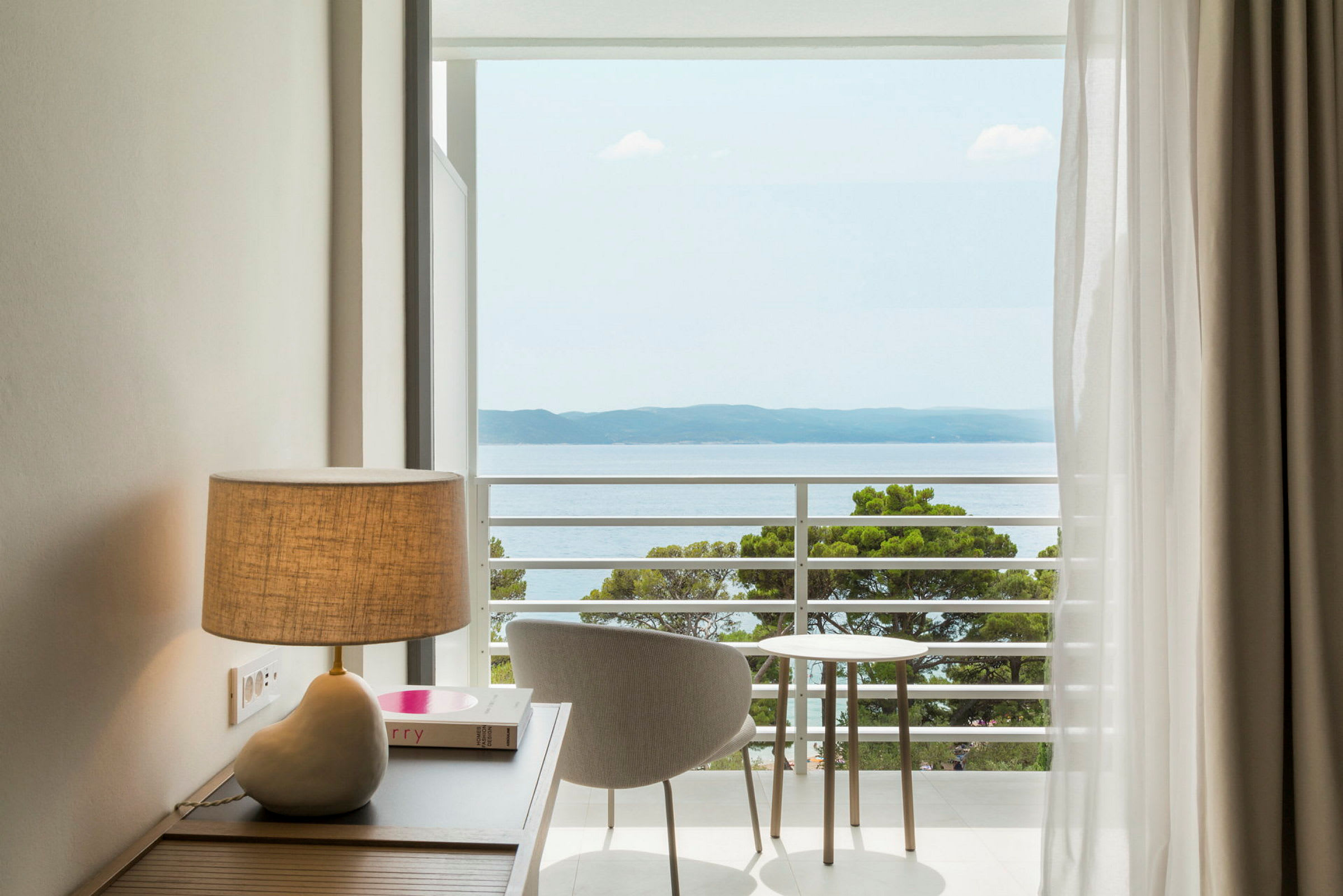 Billede af et rum med en lampe på et bord, en stol og et lille bord, der står ved en altan med udsigt over havet. (AI-genereret tekst)