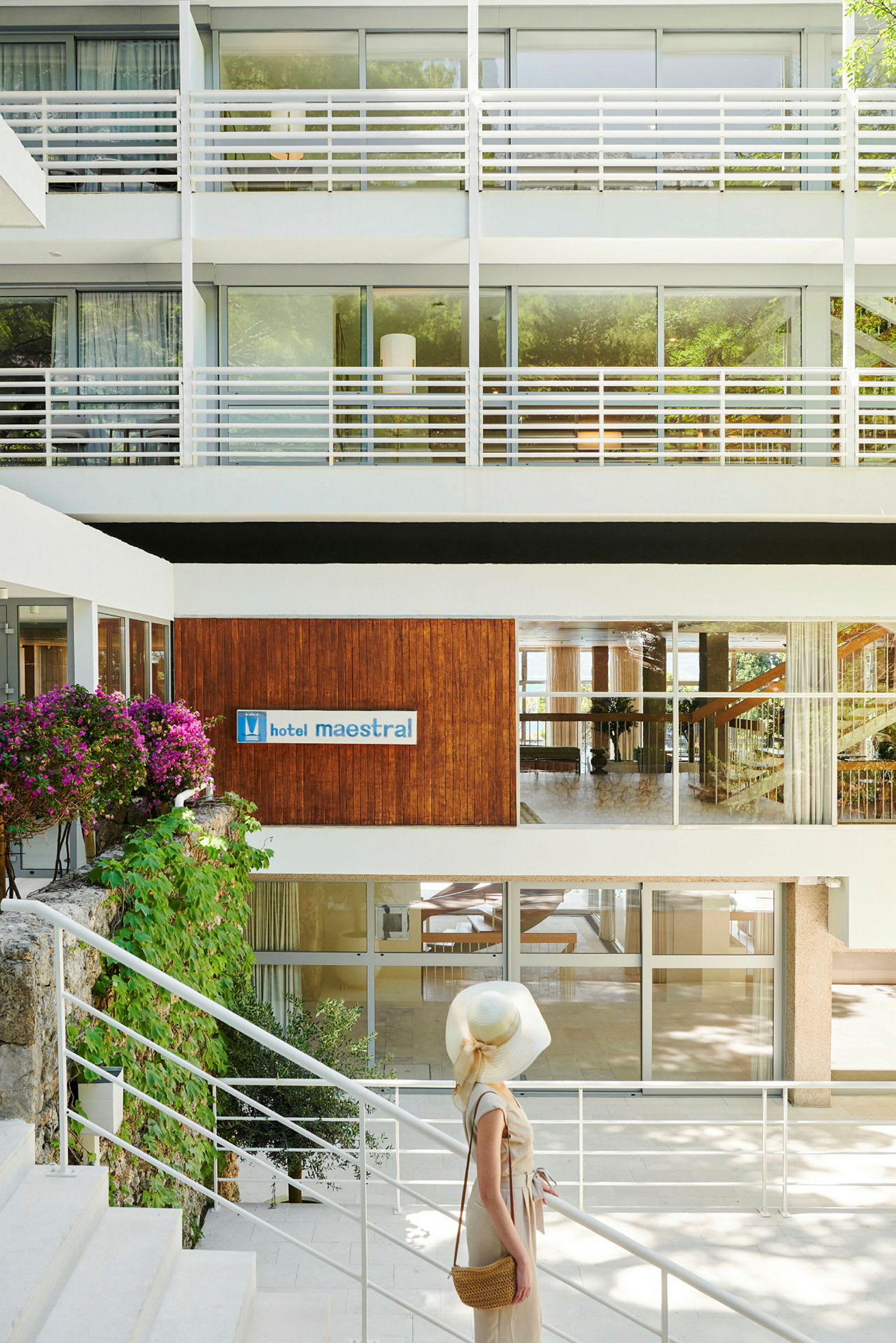 En kvinde står ved en trappe foran hotellets indgang, omgivet af moderne bygningselementer og blomster. (AI-genereret tekst)