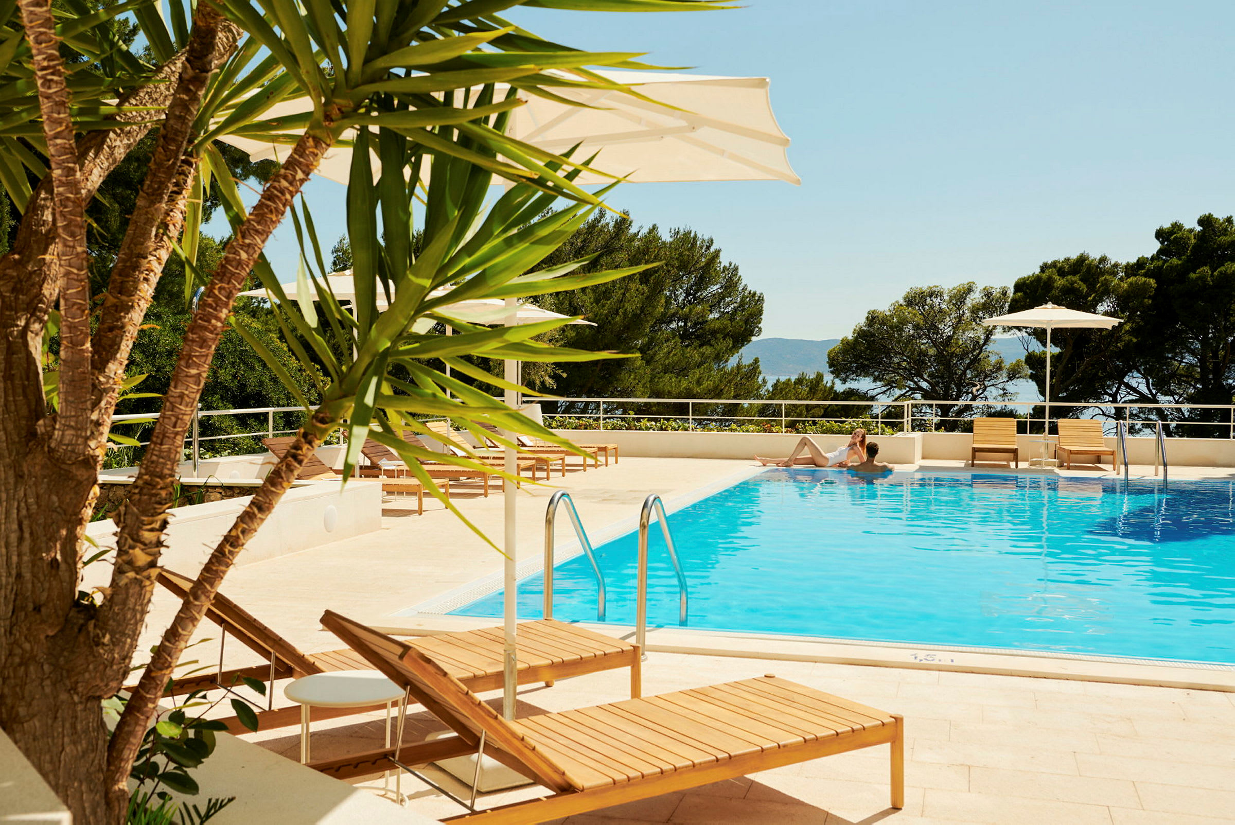 Pool med liggestole og parasoller, omgivet af træer. (AI-genereret tekst)
