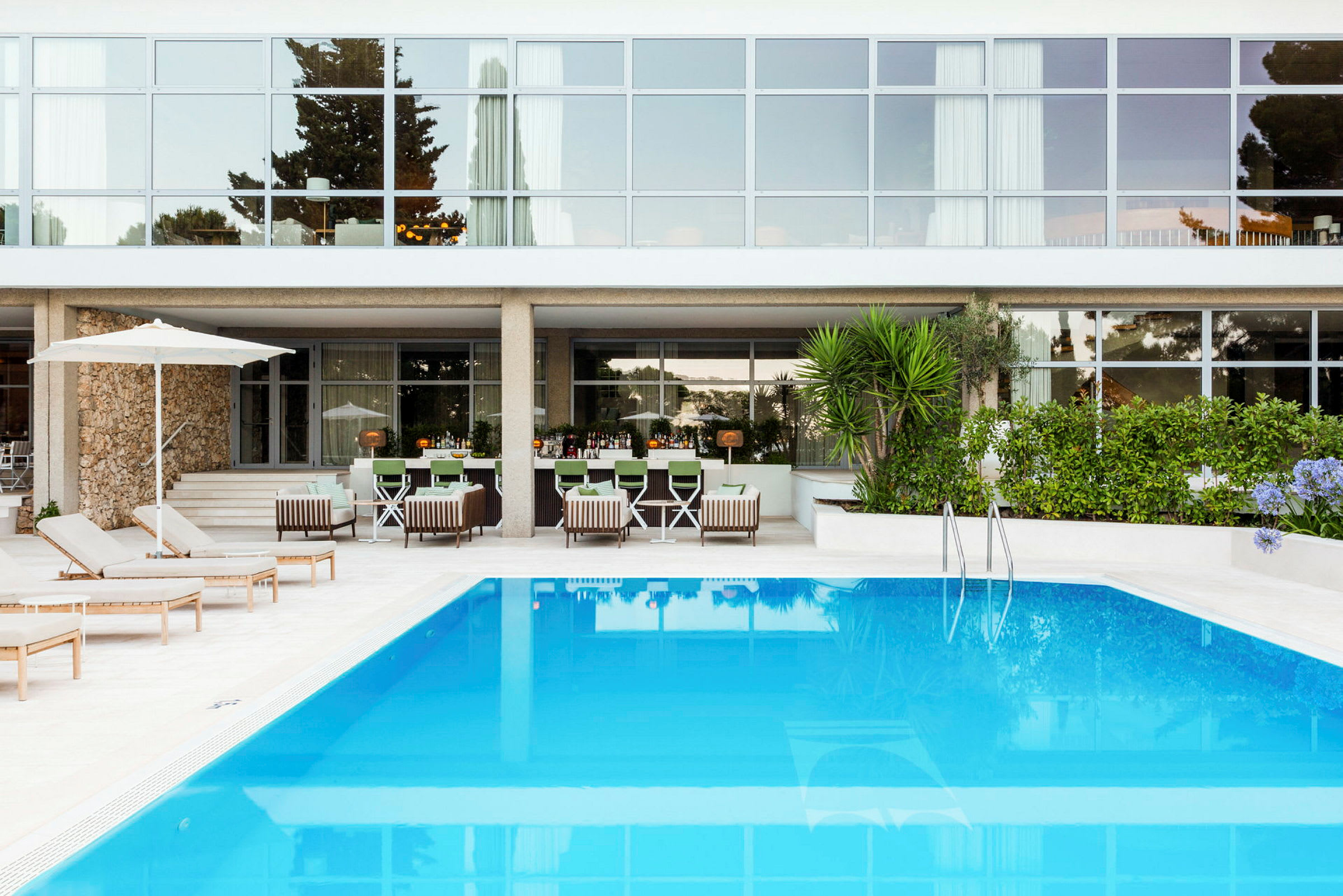 Udendørs pool foran en moderne bygning med glasfacade, omgivet af liggestole og grønne områder. (AI-genereret tekst)