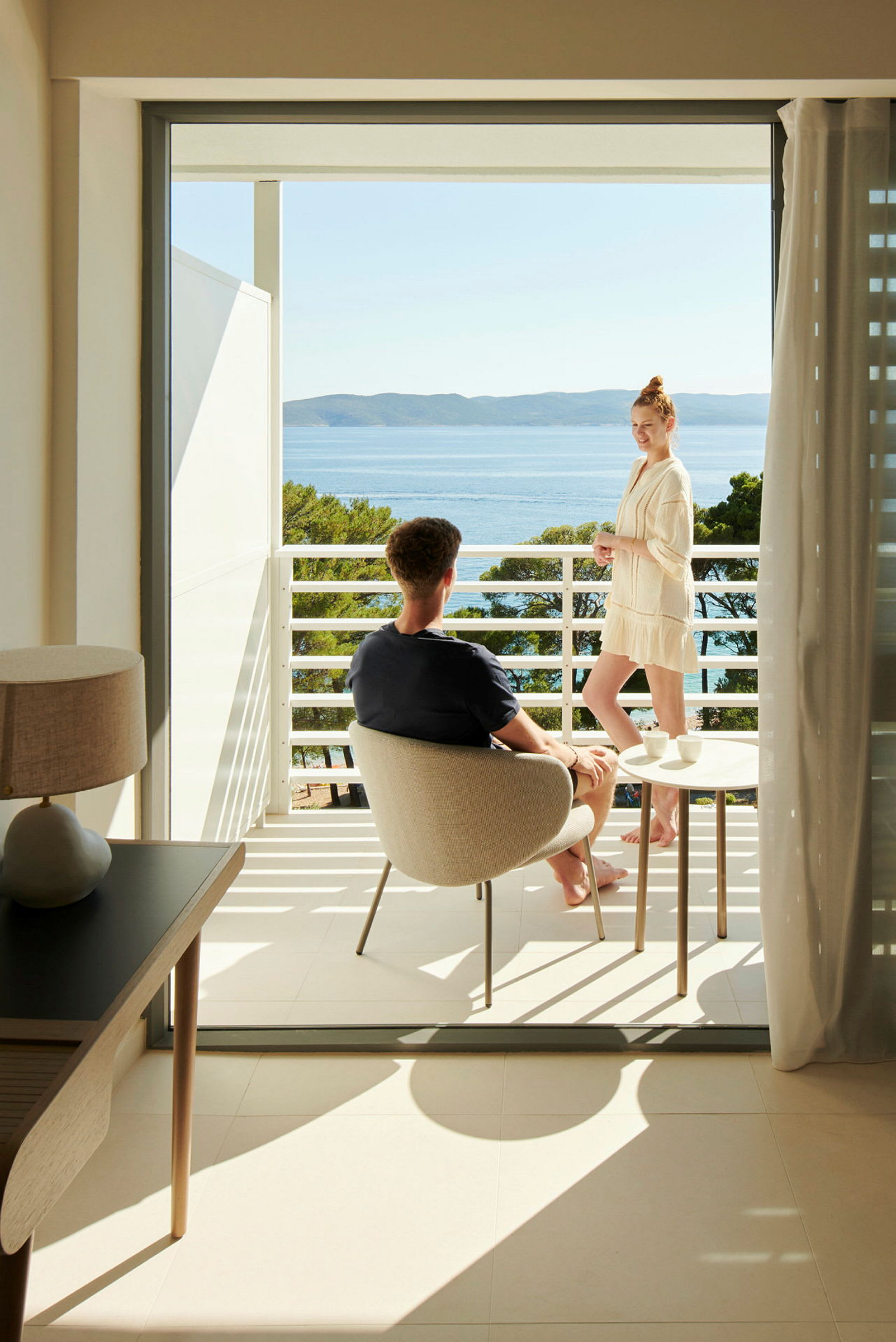 En hotelaltan med udsigt over havet og omkringliggende grønne områder. En person sidder, og en anden står ved siden af et bord. (AI-genereret tekst)